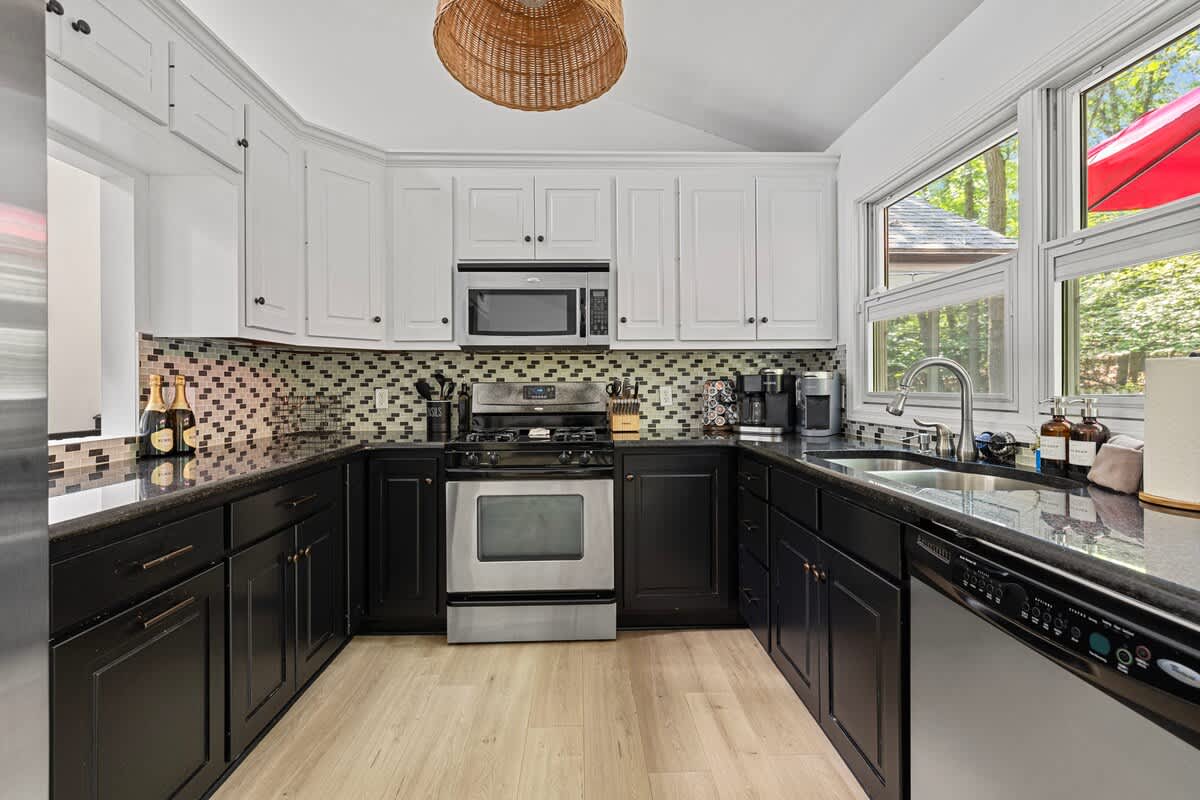 A functional kitchen with white upper and black lower cabinets, a stainless steel gas range and microwave, dark countertops, and a mosaic tile backsplash, illuminated by natural light from a large window.