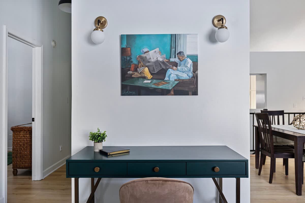 Dedicated workspace with a dark green desk, a light brown velvet chair, and brass wall sconces, featuring a colorful painting above the desk and light wood flooring.