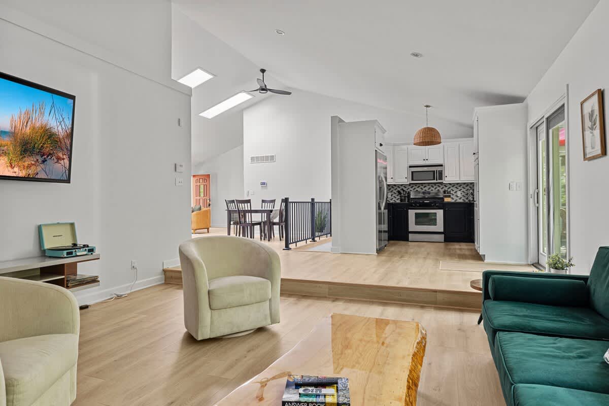 A bright open-concept living space featuring a large wall-mounted TV, two light-colored swivel armchairs, and a dark green sofa, with a raised dining area and a modern kitchen visible in the background.