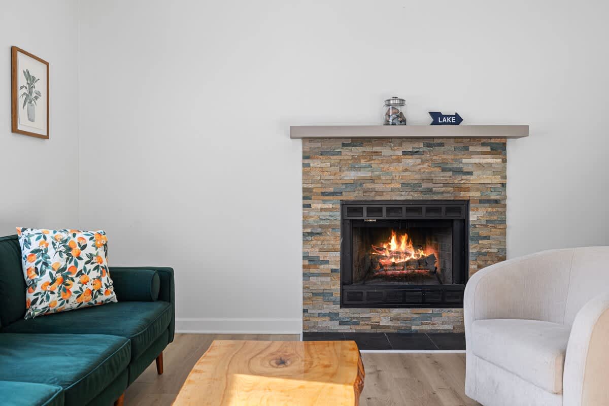 A close-up of the living room's stone-clad fireplace, featuring a warm fire burning within, a simple mantelpiece adorned with a glass jar and a "LAKE" sign, and light wood flooring.