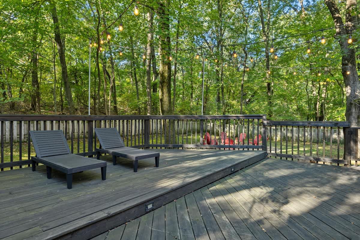 A spacious dark wooden deck with two modern dark gray lounge chairs, illuminated by string lights strung between tall trees, offering a view of a fire pit area in the distance.