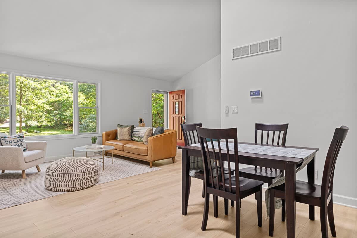 A bright open-concept space featuring a dark wood dining table with four chairs in the foreground, leading into a living area with a light brown leather sofa, a patterned ottoman, and large windows overlooking lush greenery.