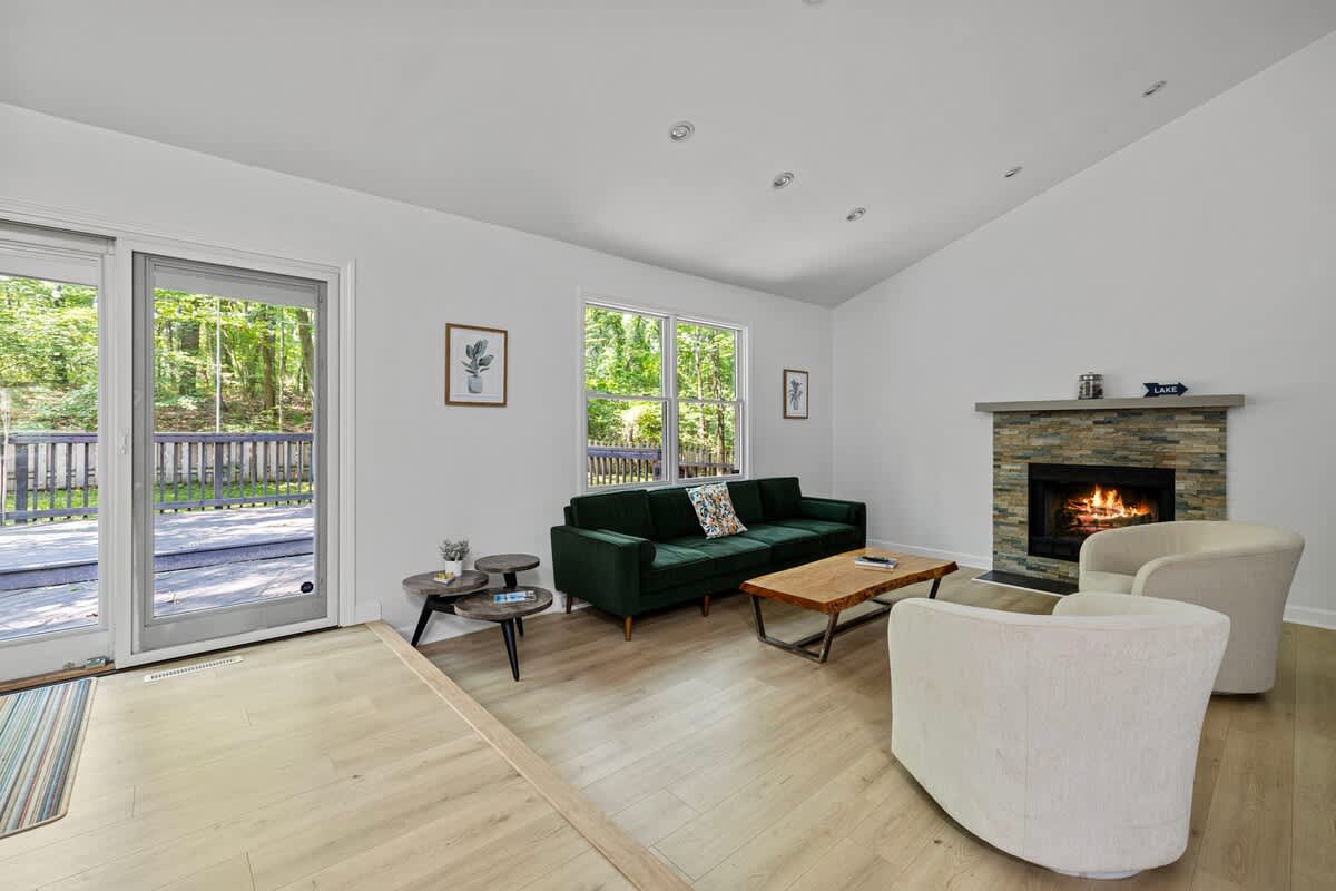 A cozy living room with a dark green velvet sofa, two light-colored armchairs, and a stone-clad fireplace with a burning fire, all set against light wood floors and large glass doors opening to an outdoor deck.