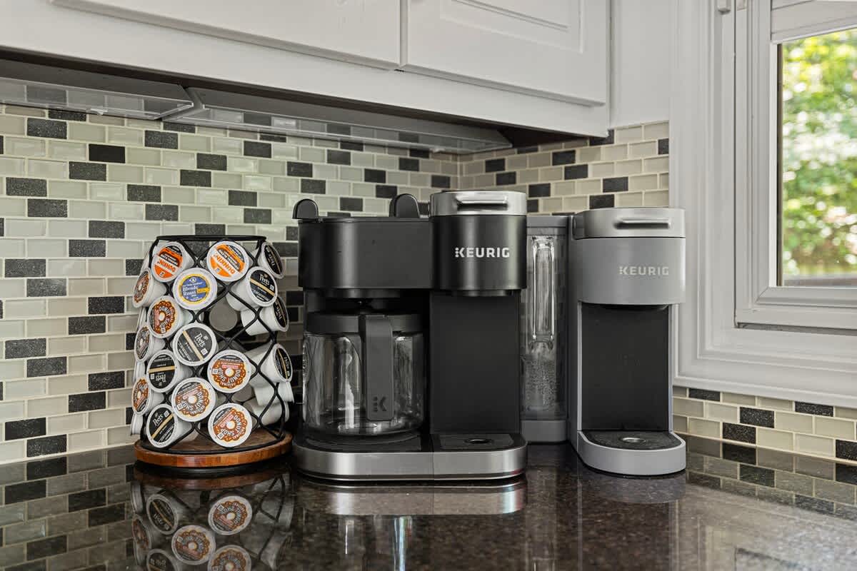 A modern kitchen counter features two black and silver Keurig coffee makers and a black metal K-cup holder filled with various coffee pods, set against a mosaic tile backsplash and dark granite countertop.