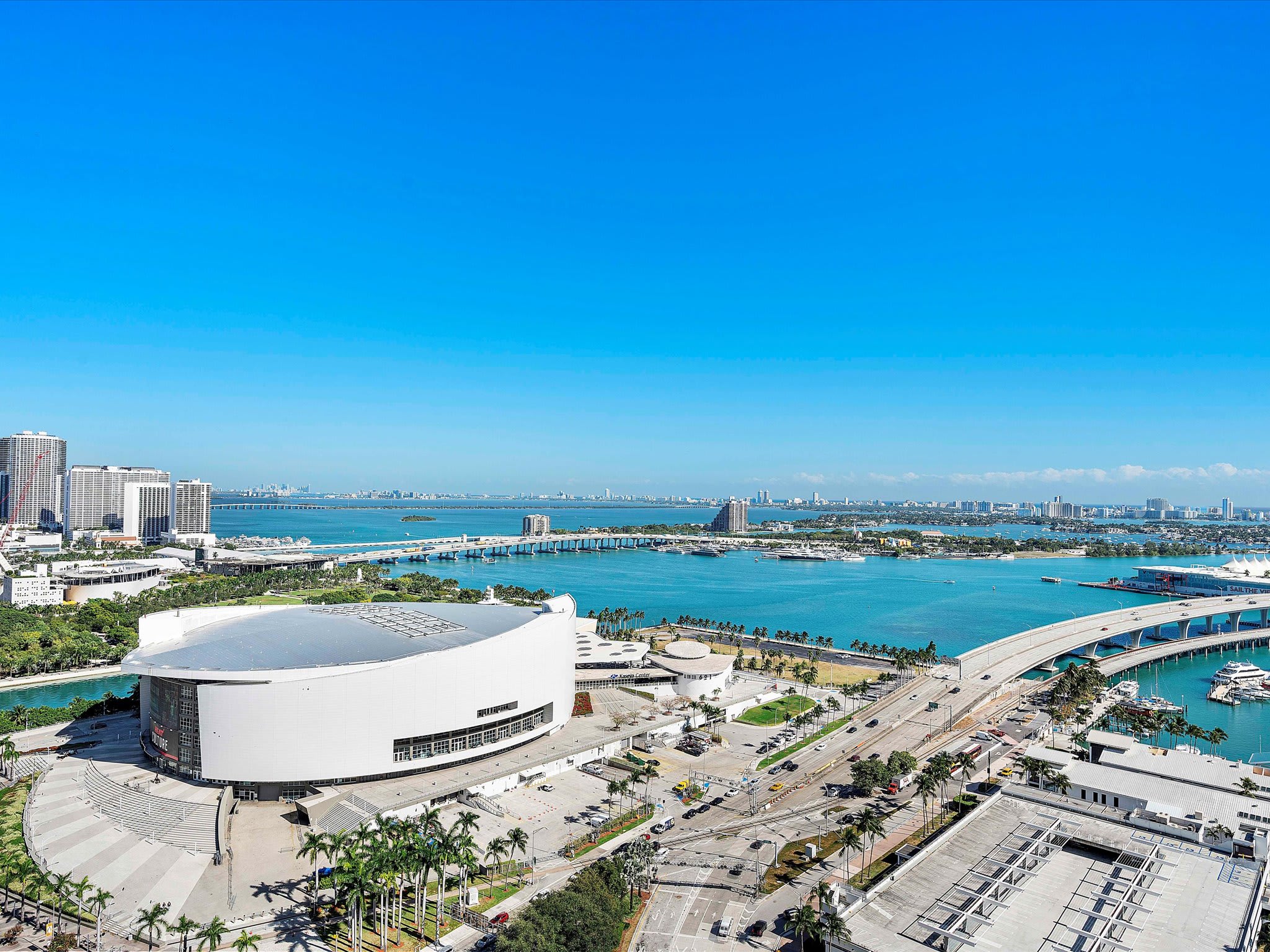 An expansive aerial view showcases a vibrant city skyline with numerous high-rise buildings, a large stadium, and bridges spanning across a wide, clear blue bay. The scene is bathed in bright sunlight under a clear sky, highlighting the urban landscape and waterfront.