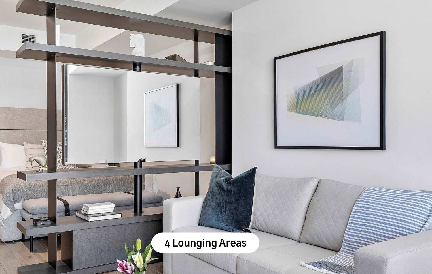 Bright living area featuring a light-colored sofa with a dark blue accent pillow, an abstract framed artwork, and a modern dark wood shelving unit serving as a room divider.