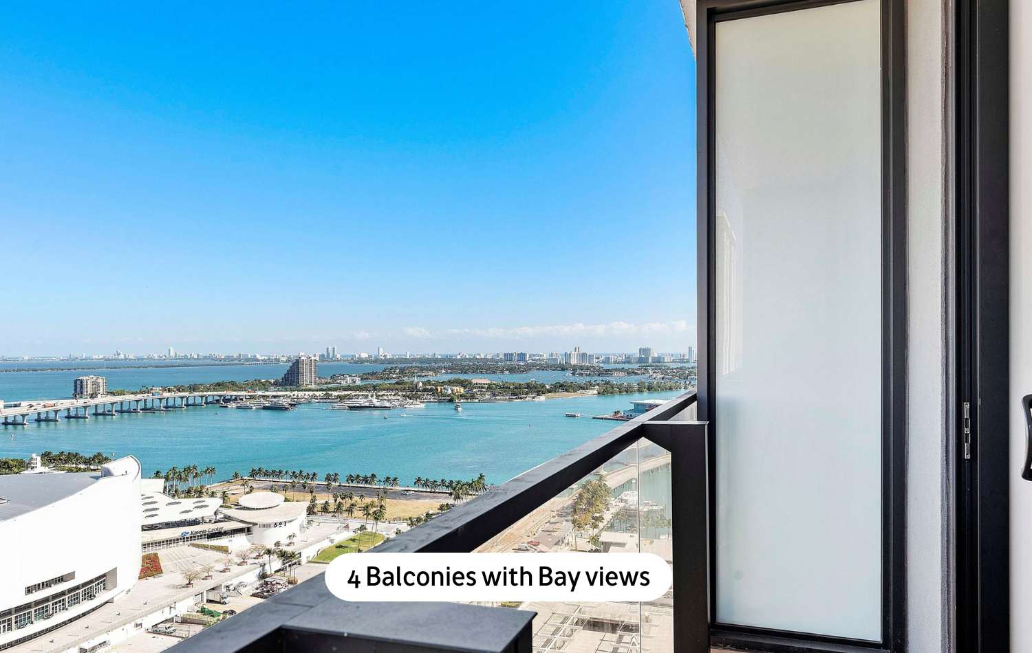 Private balcony with a clear glass railing offering breathtaking panoramic bay views, a distant city skyline, and a bright blue sky, perfect for outdoor relaxation.