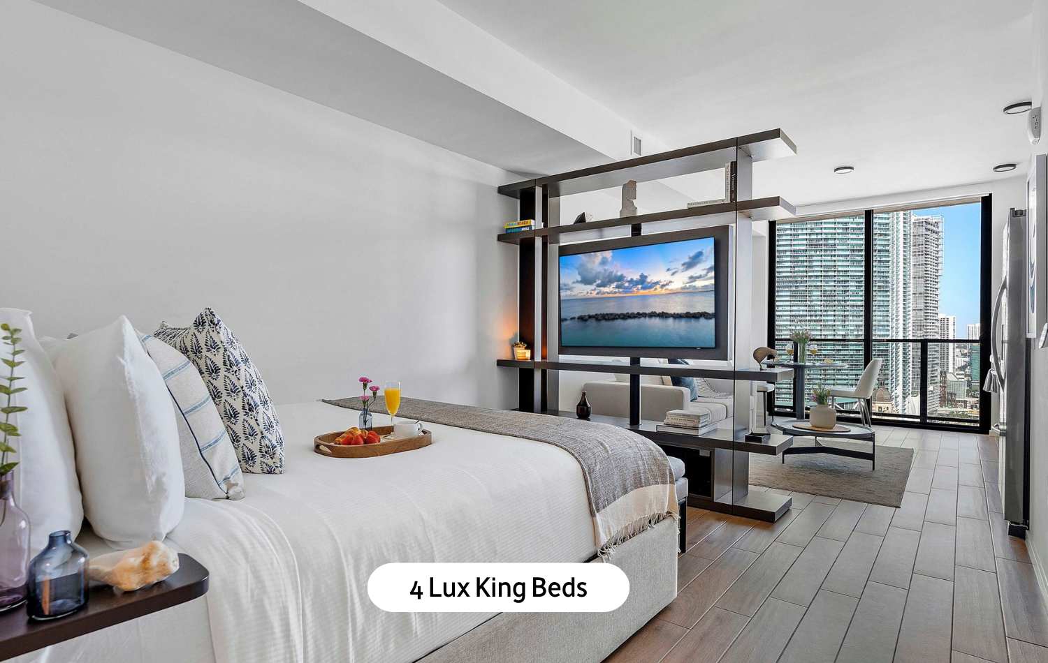 Modern bedroom featuring a luxurious king-sized bed with a breakfast tray, a flat-screen TV integrated into a stylish dark wood room divider, and a seating area with expansive city views through large windows.