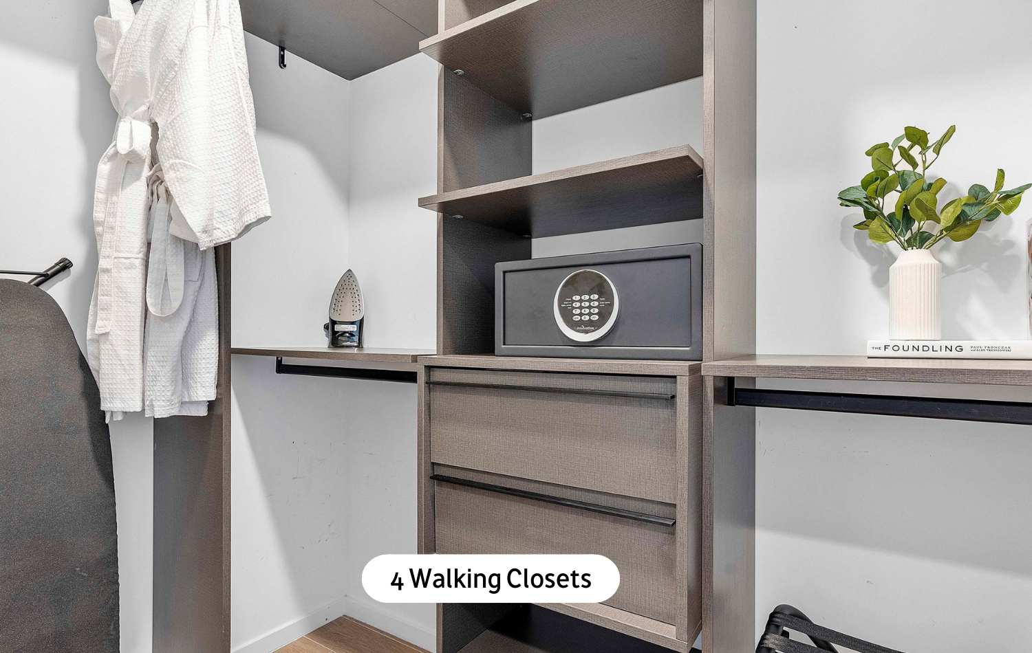 Organized walk-in closet featuring built-in dark wood shelving, drawers, and hanging rods, complete with a personal safe, iron, and a white bathrobe.