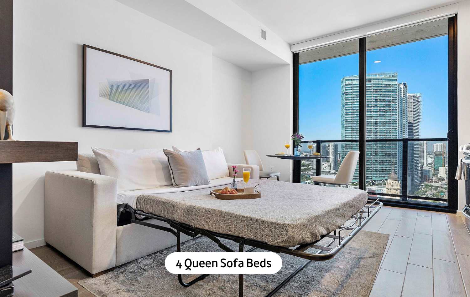 Bright studio space featuring a queen sofa bed, a small dining table for two, and an abstract artwork, all set against a backdrop of large windows with a vibrant city view.
