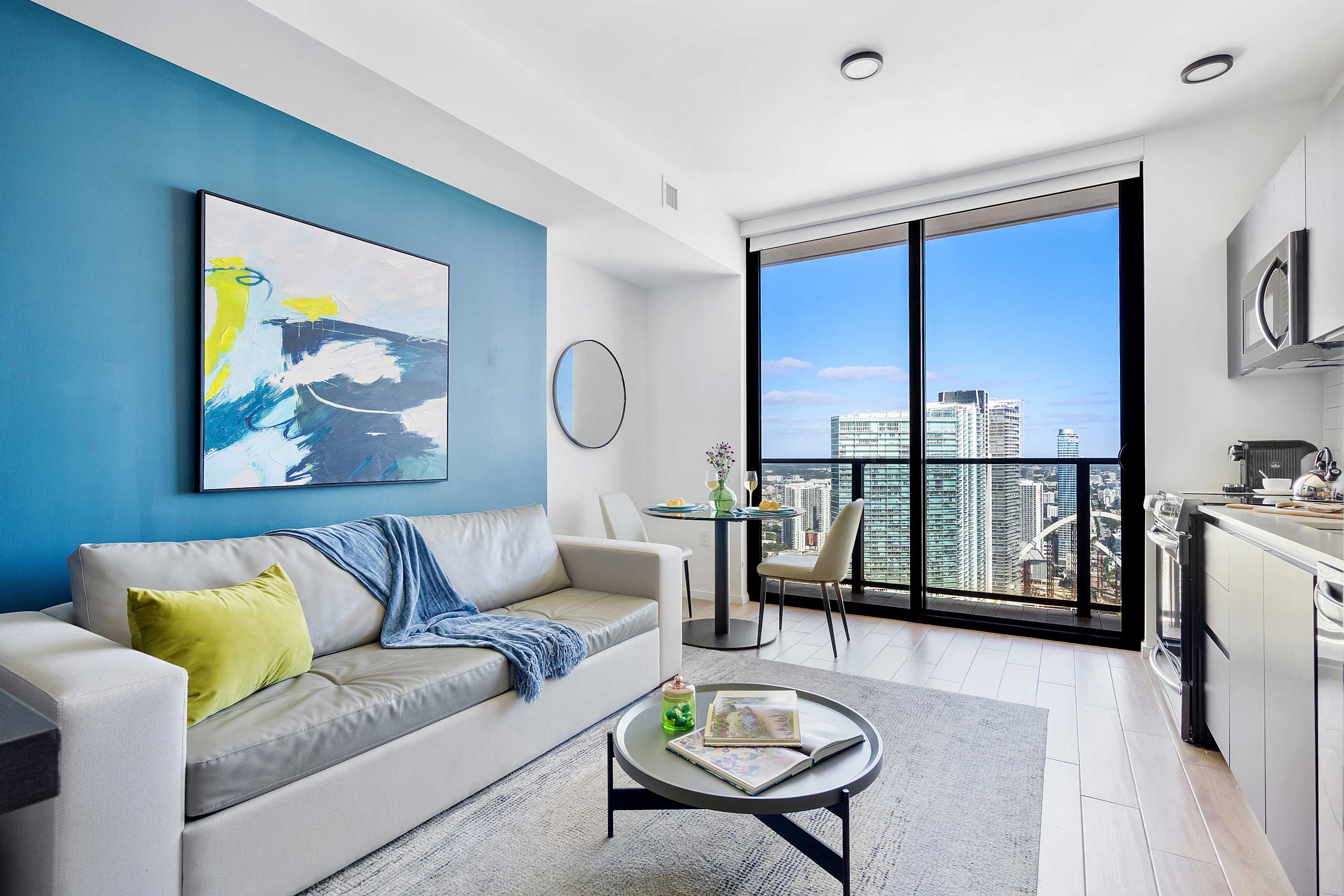 Bright living area of a studio apartment featuring a light gray sofa, a round dining table, and a large floor-to-ceiling window offering city views. A blue accent wall with abstract art adds a pop of color.