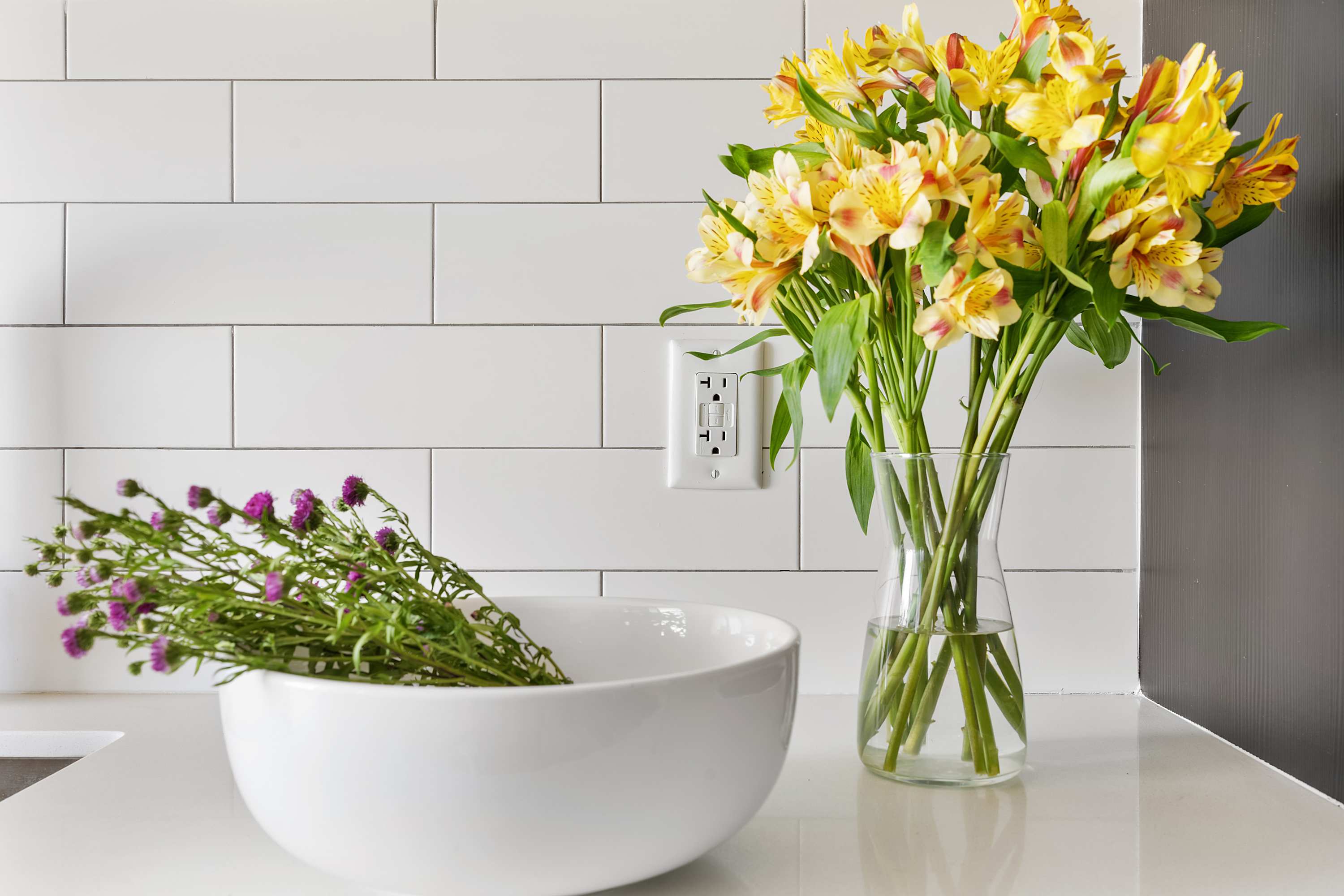 Bright kitchen counter with a white subway tile backsplash, showcasing a large glass vase filled with vibrant yellow and orange alstroemeria flowers next to a white bowl containing purple wildflowers.