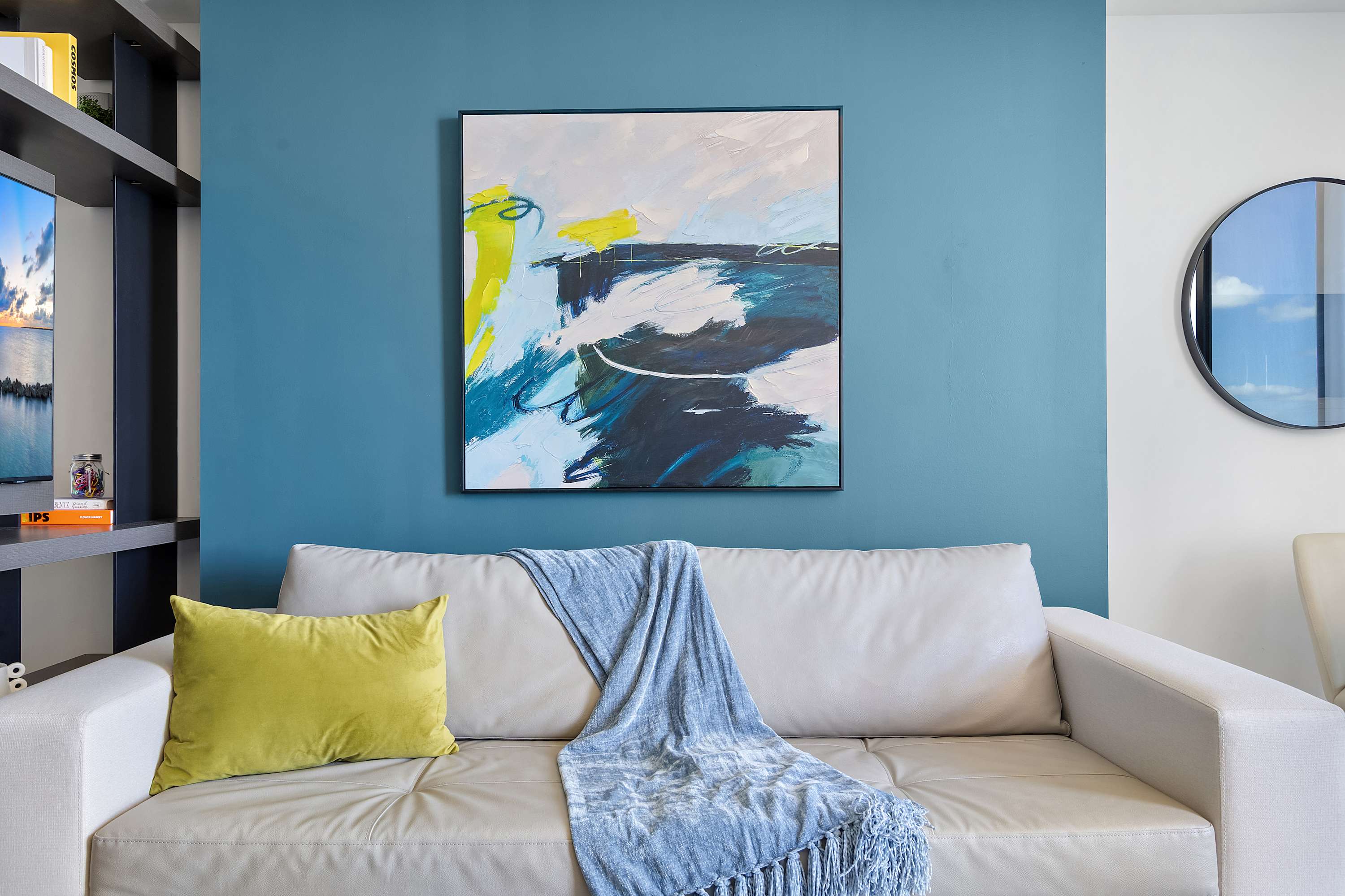 Close-up of a light gray sofa with a chartreuse pillow and blue throw, set against a vibrant blue accent wall adorned with a large abstract painting.