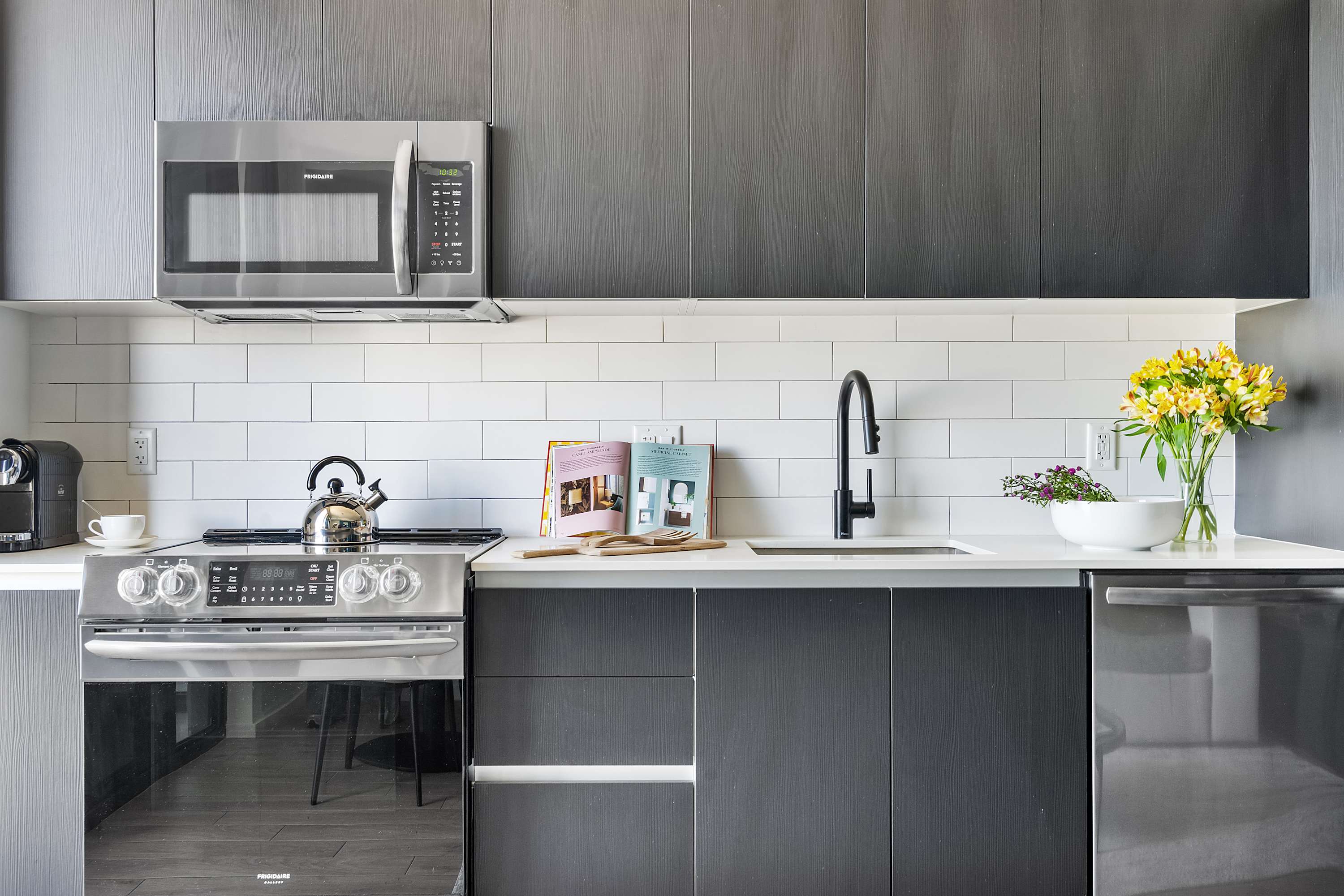 Sleek, modern kitchen with dark gray flat-panel cabinetry, white quartz countertops, and a white subway tile backsplash. Stainless steel appliances include a microwave, gas range, and a black faucet over the sink.