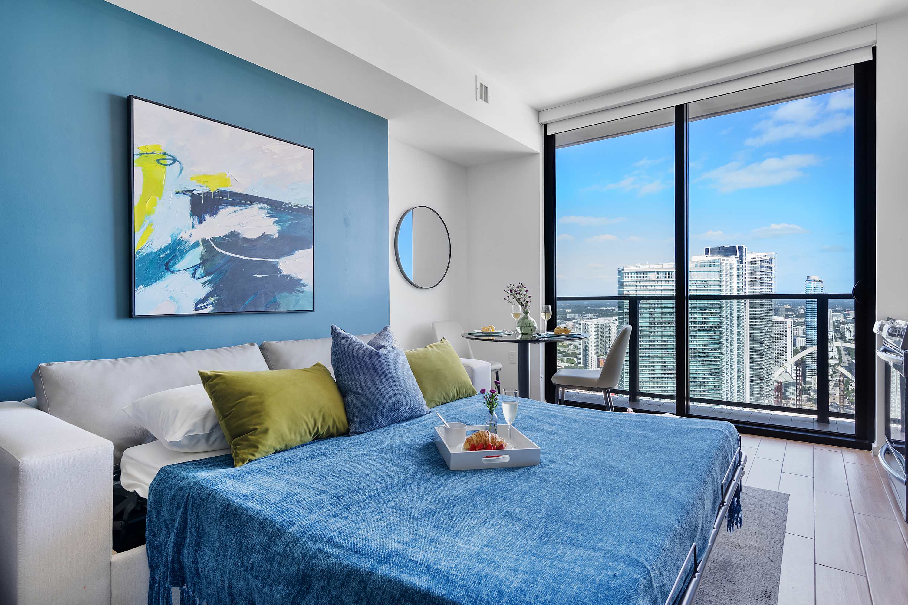 Studio apartment with a sofa bed pulled out, covered in a blue throw, facing a large window with city views. A blue accent wall with abstract art and a small dining table are also visible.