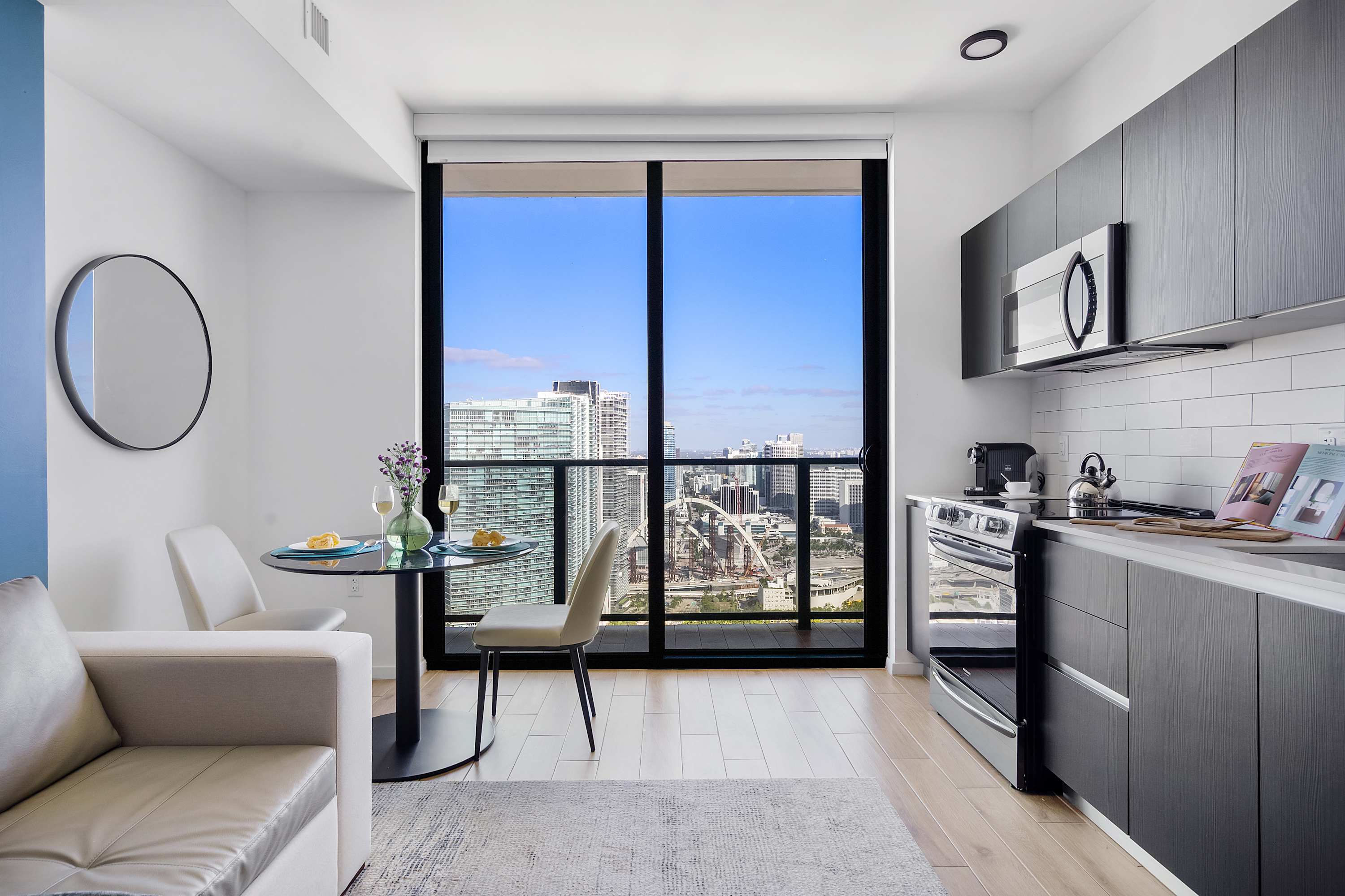 Modern open-concept studio living space featuring a compact kitchen, a small dining area, and a sofa, all with light wood floors and a large window offering panoramic city views.