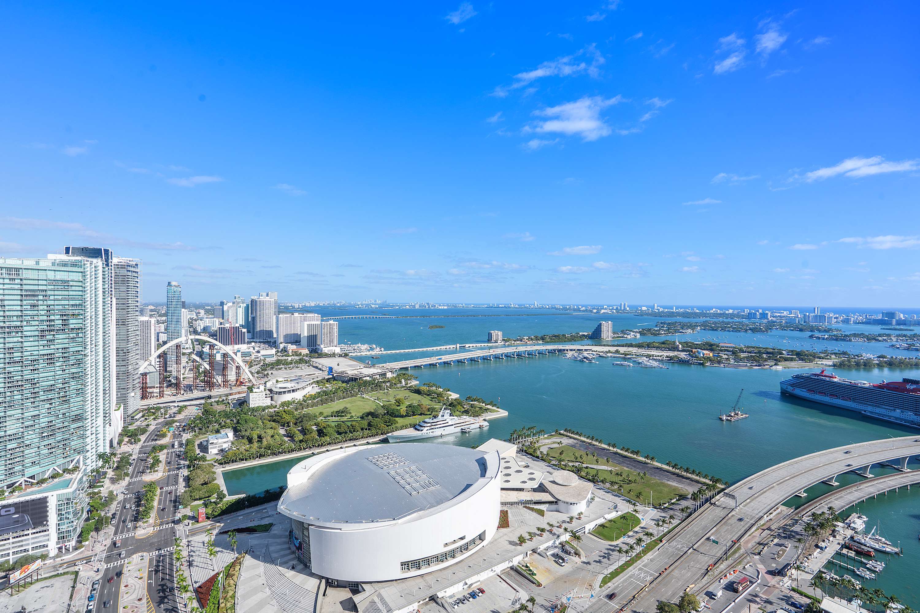 Expansive aerial view showcasing a bustling city waterfront with a large stadium, numerous high-rise buildings, and bridges spanning across the bay, all under a bright blue sky.