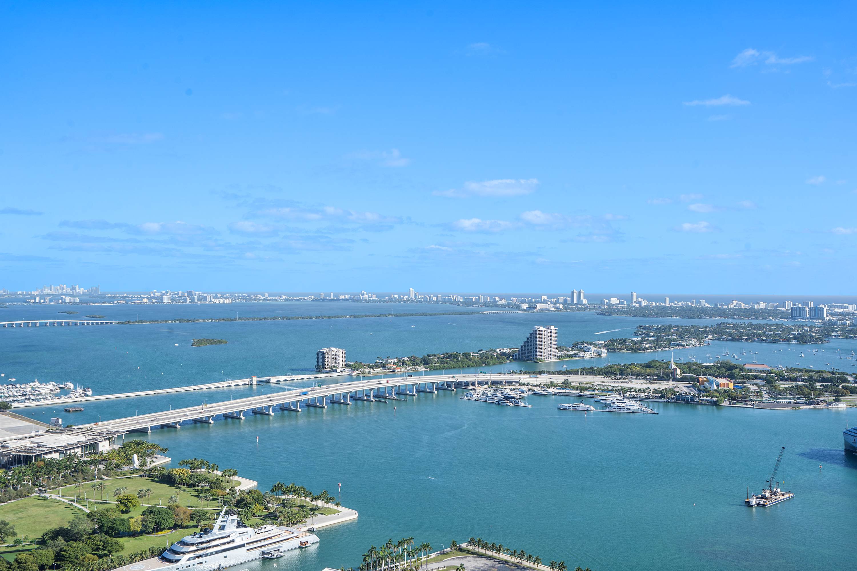 Wide panoramic view of a vibrant bay area with multiple bridges, islands, and boats, surrounded by a distant city skyline under a bright blue sky with scattered clouds.