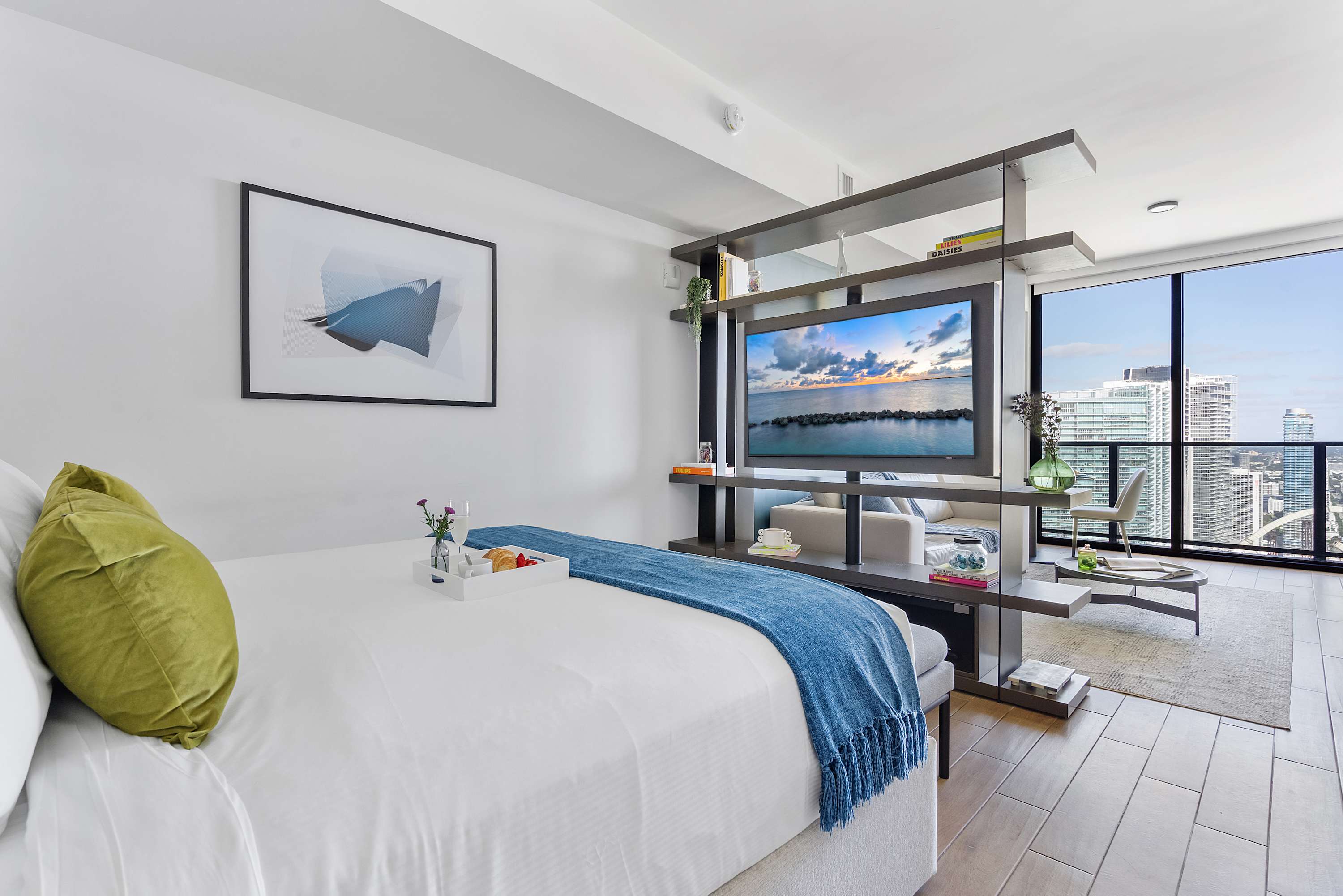Studio bedroom with a king bed, light wood floors, and a modern dark shelving unit acting as a room divider, featuring a mounted TV and offering a glimpse of the living area and city views beyond.