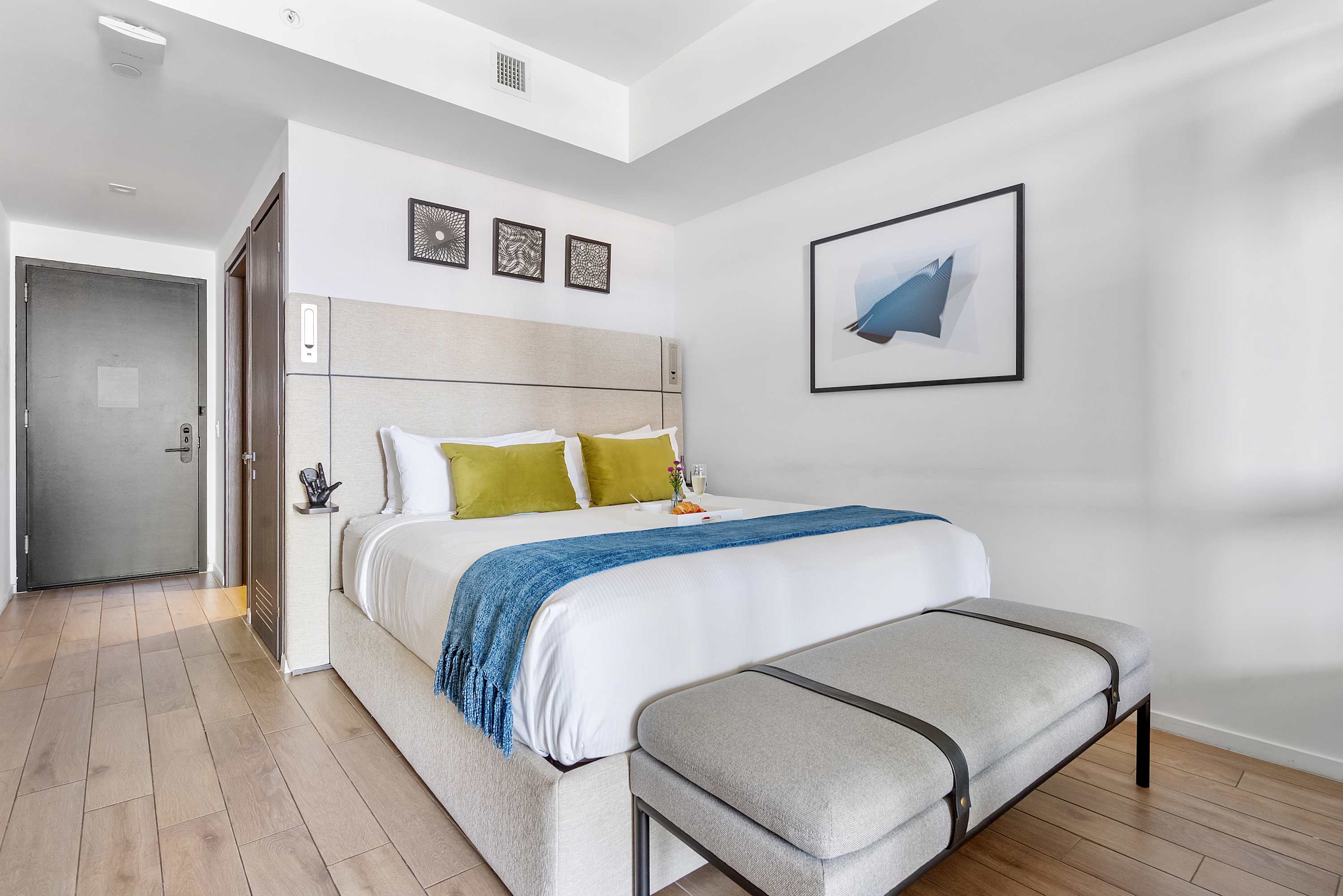 Elegant bedroom featuring a king-sized bed with a light grey upholstered headboard, crisp white bedding, green accent pillows, and a blue throw, complemented by light wood floors and modern framed artwork.