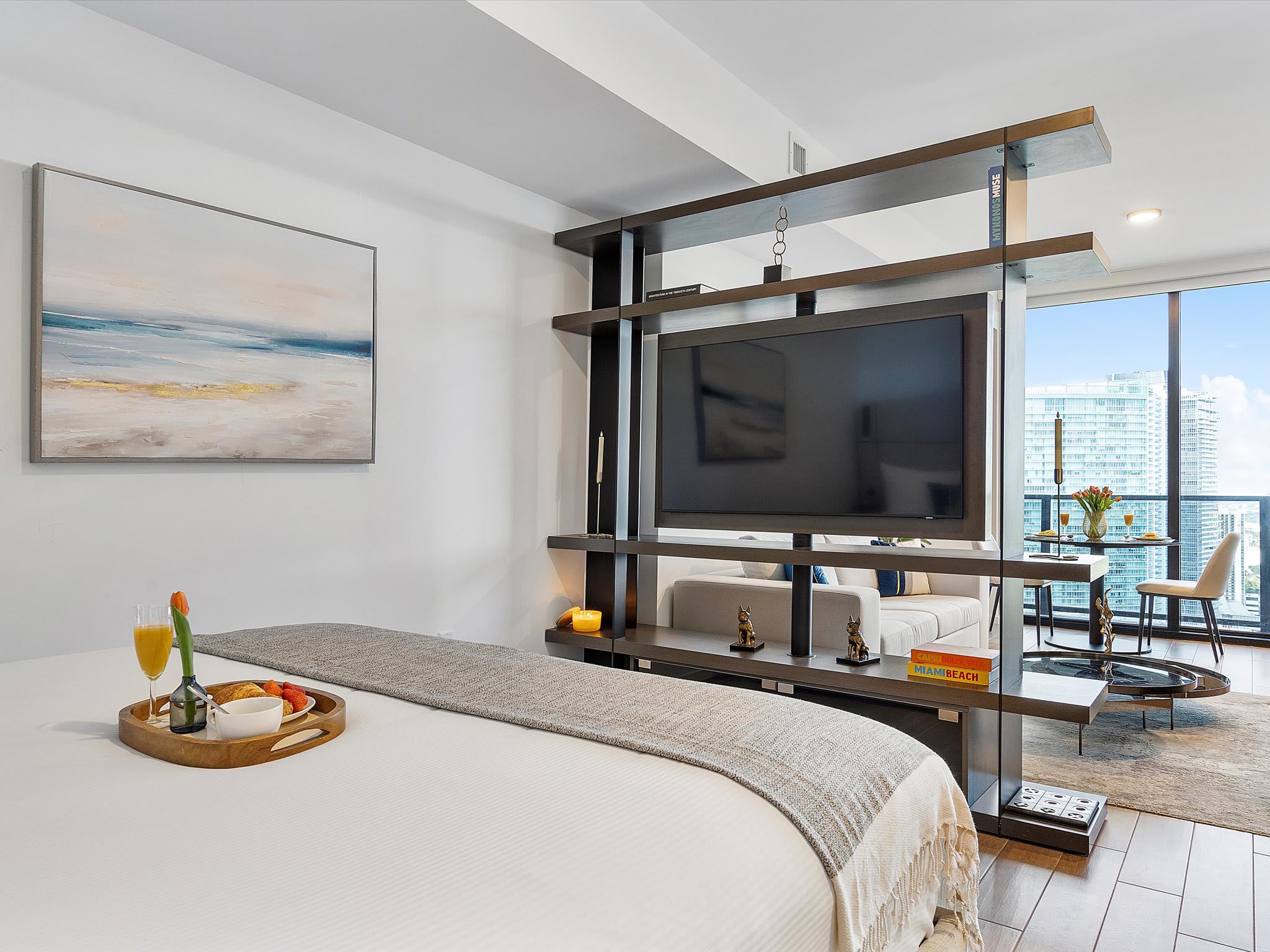 Studio bedroom area featuring a large bed with a grey throw, separated from the living space by a dark wood shelving unit holding a flat-screen TV. A modern abstract painting hangs on the white wall, and a breakfast tray is on the bed.