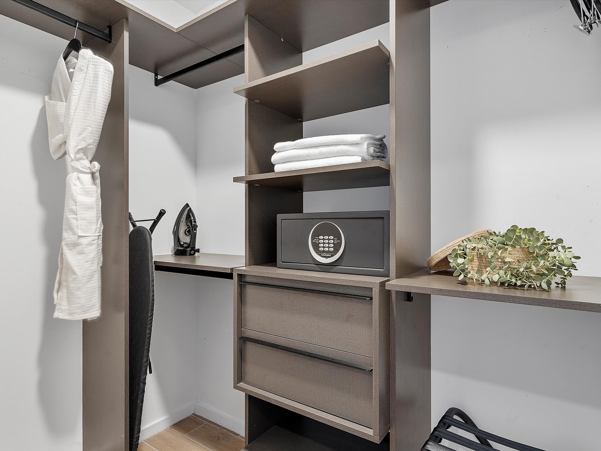 Organized walk-in closet with dark wood shelving and hanging space, featuring a white bathrobe, iron, ironing board, and a digital safe. Freshly folded white towels are neatly stacked on a shelf.