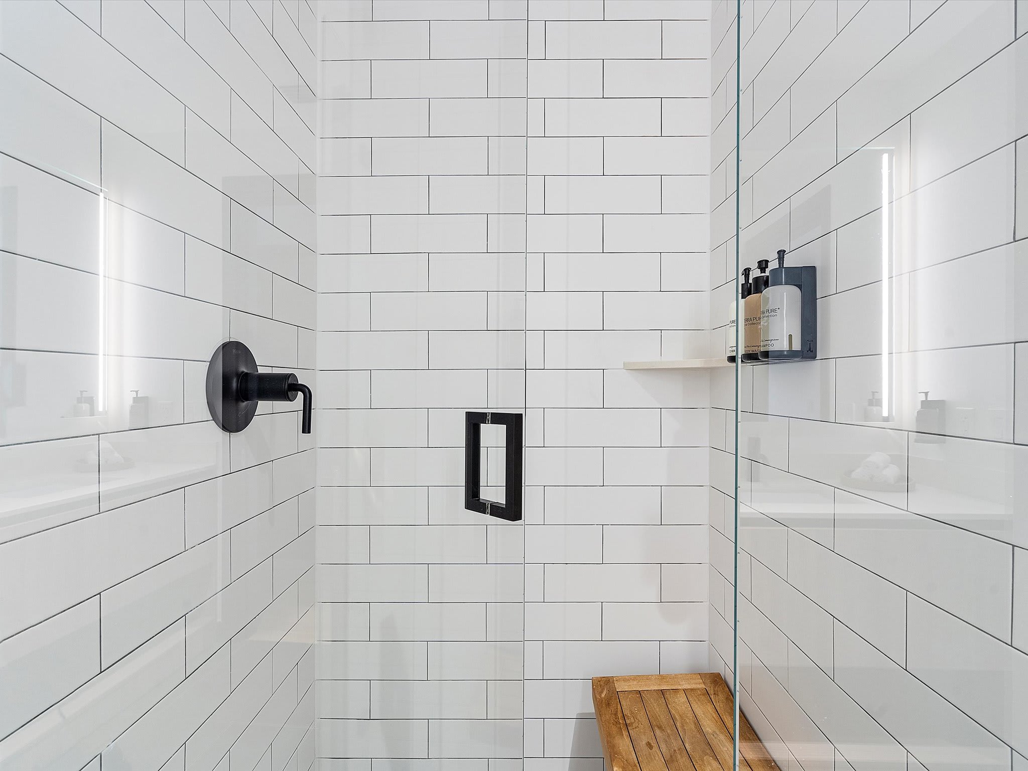 Modern walk-in shower with white subway tiles, a black matte shower head, and a built-in black niche. A glass partition separates the shower, revealing a wooden bench and toiletries on a shelf.
