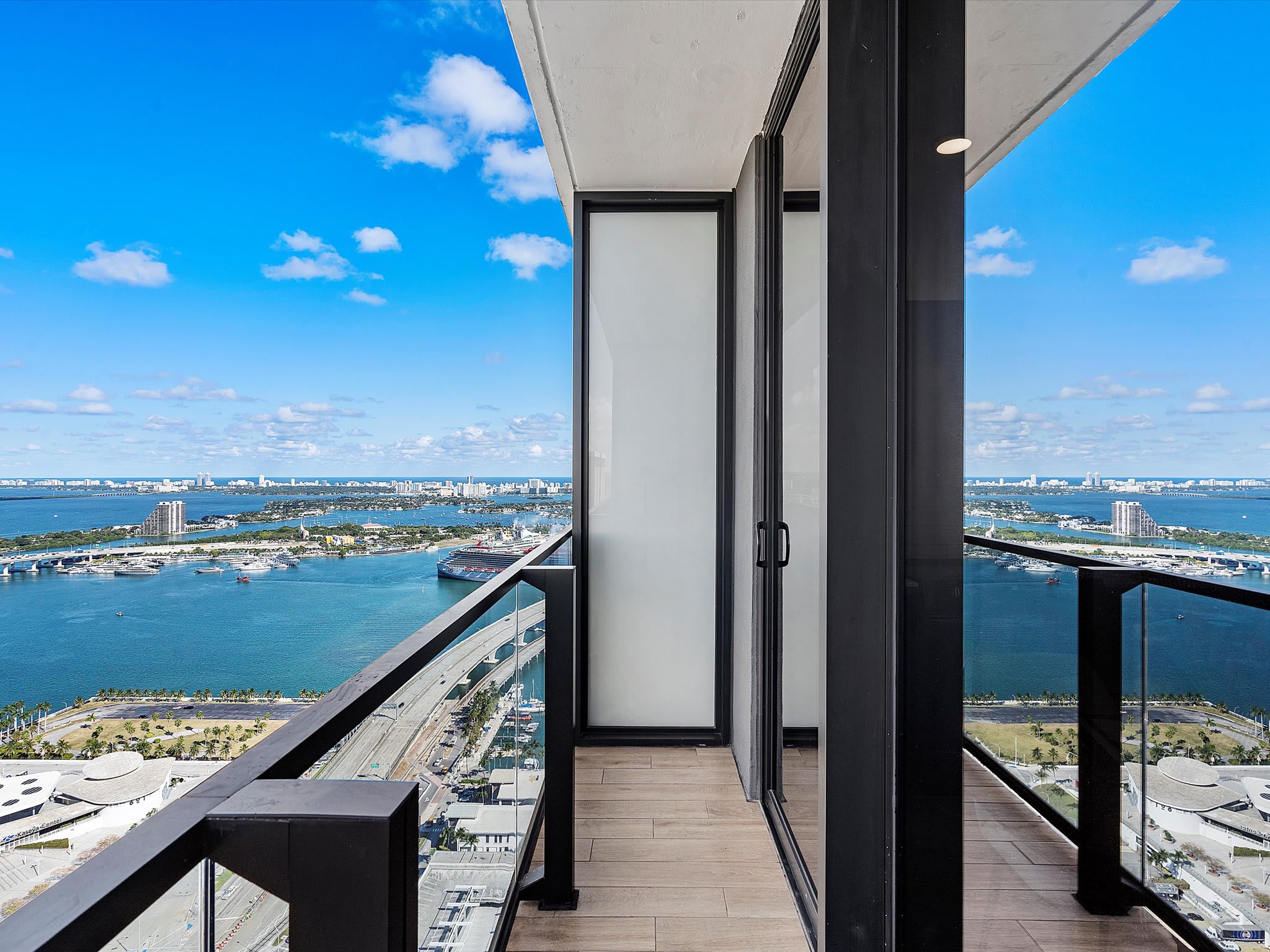Spacious balcony with glass railings offering panoramic views of the bay, city skyline, and distant islands under a clear blue sky. Sliding glass doors lead back into the interior space.