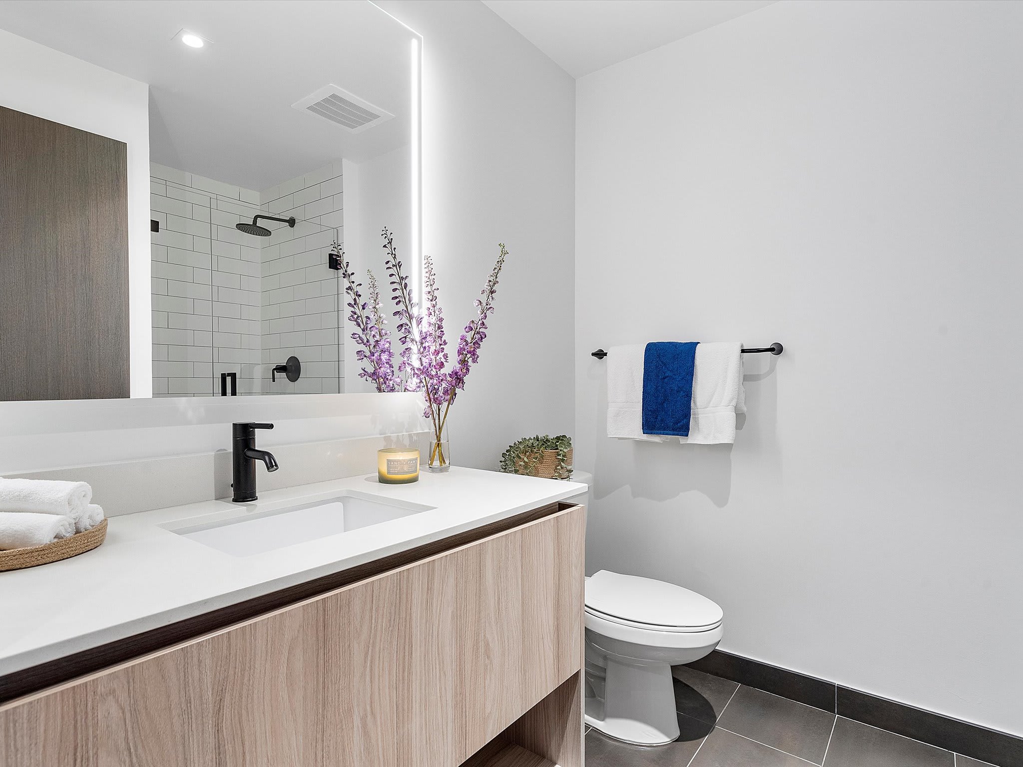 Contemporary bathroom with a light wood vanity, white rectangular sink, black faucet, and a large illuminated mirror. A white toilet is next to the vanity, and a tiled shower with a black fixture is visible in the background.