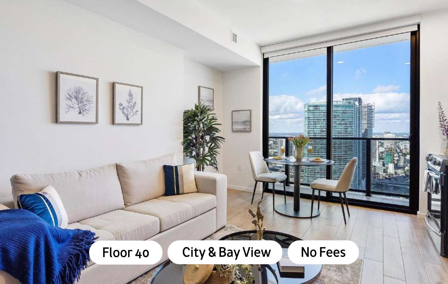 Bright, modern living space with a light beige sofa, a round glass dining table for two, and floor-to-ceiling windows offering expansive city and bay views. Light wood-look flooring and minimalist decor complete the contemporary feel.