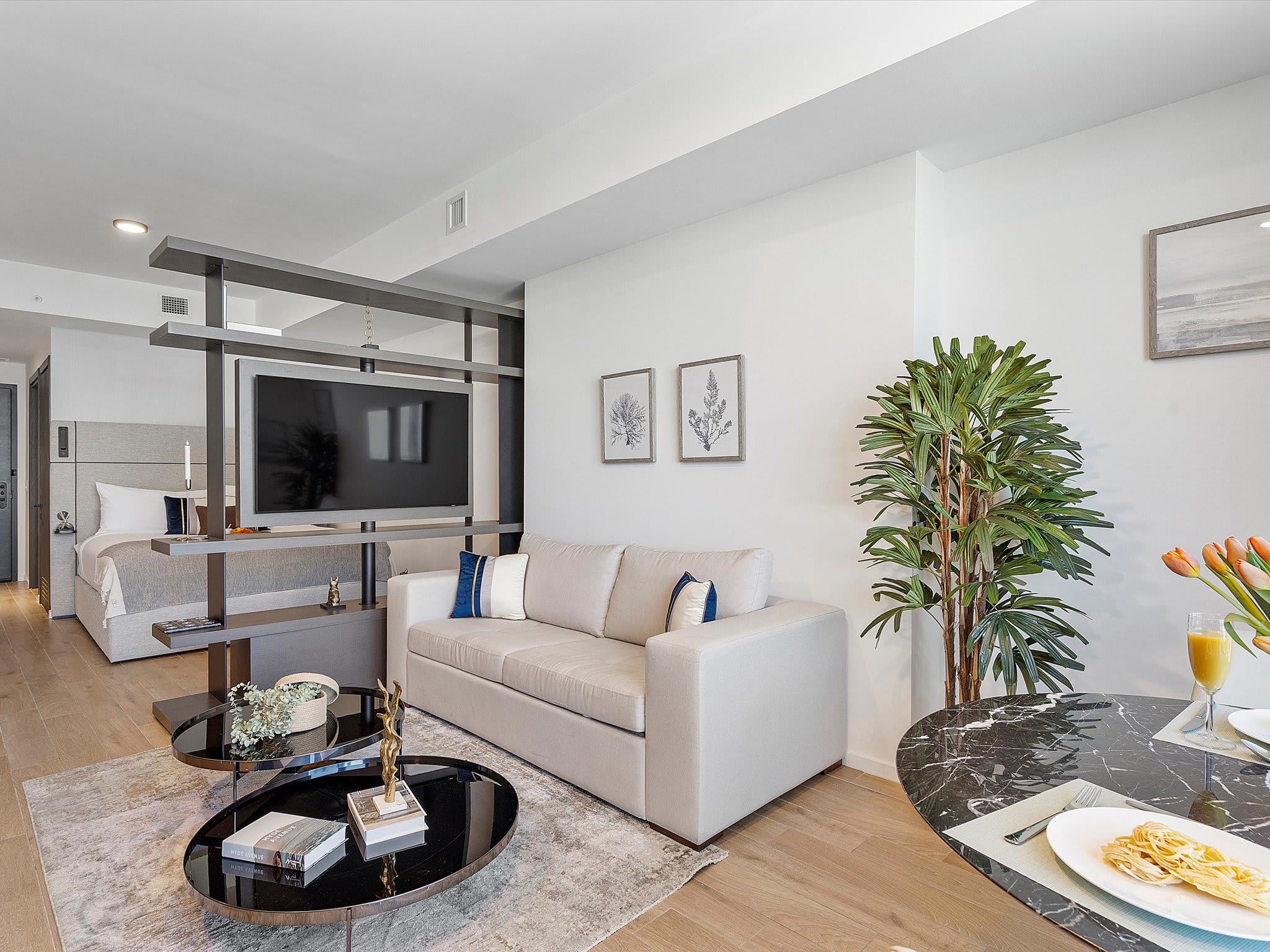 Contemporary studio living space with a light beige sofa, a dark marble-top dining table, and a multi-tiered coffee table on an area rug. A dark shelving unit partially separates the living area from the bedroom, with large windows offering city views.