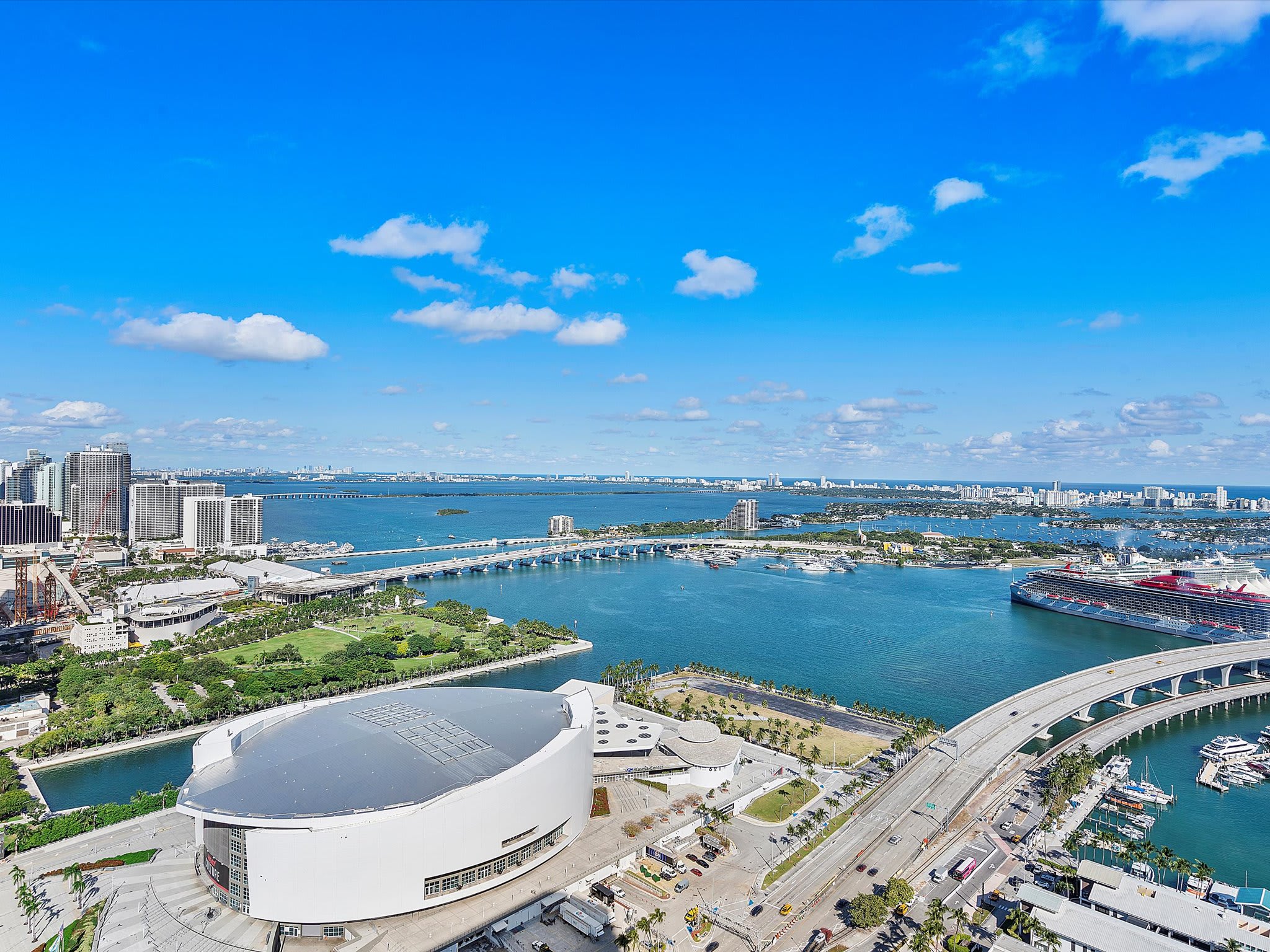 Expansive aerial view showcasing a vibrant city skyline alongside a large bay with multiple bridges and a cruise ship docked. A prominent sports arena is visible in the foreground.