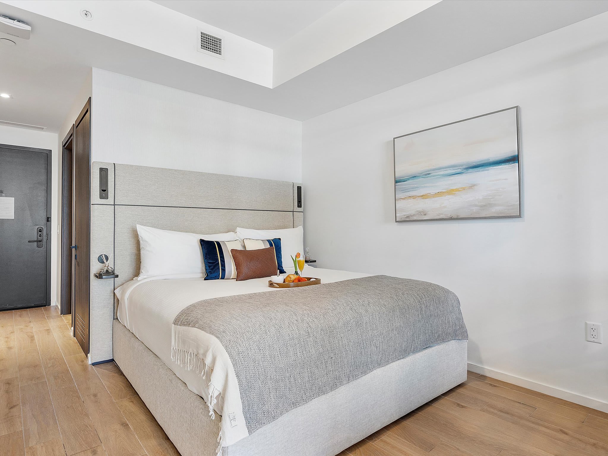 Modern studio bedroom featuring a queen bed with a grey upholstered headboard, light wood flooring, and a contemporary abstract painting on the white wall. A tray with drinks and fruit rests on the bed.