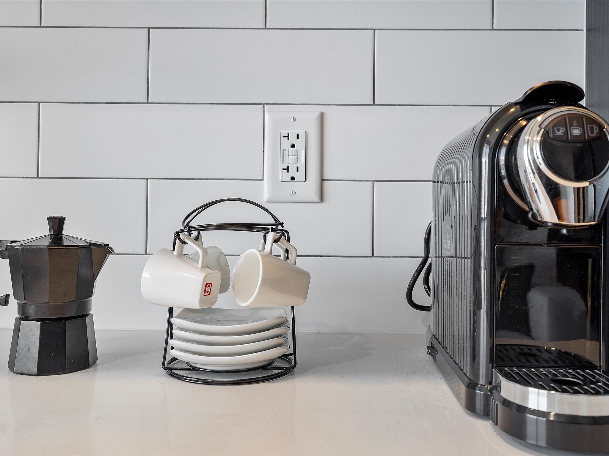 Kitchen countertop featuring a black espresso machine, a traditional Moka pot, and a stand with white coffee cups and saucers. A white electrical outlet is visible against the white subway tile backsplash.