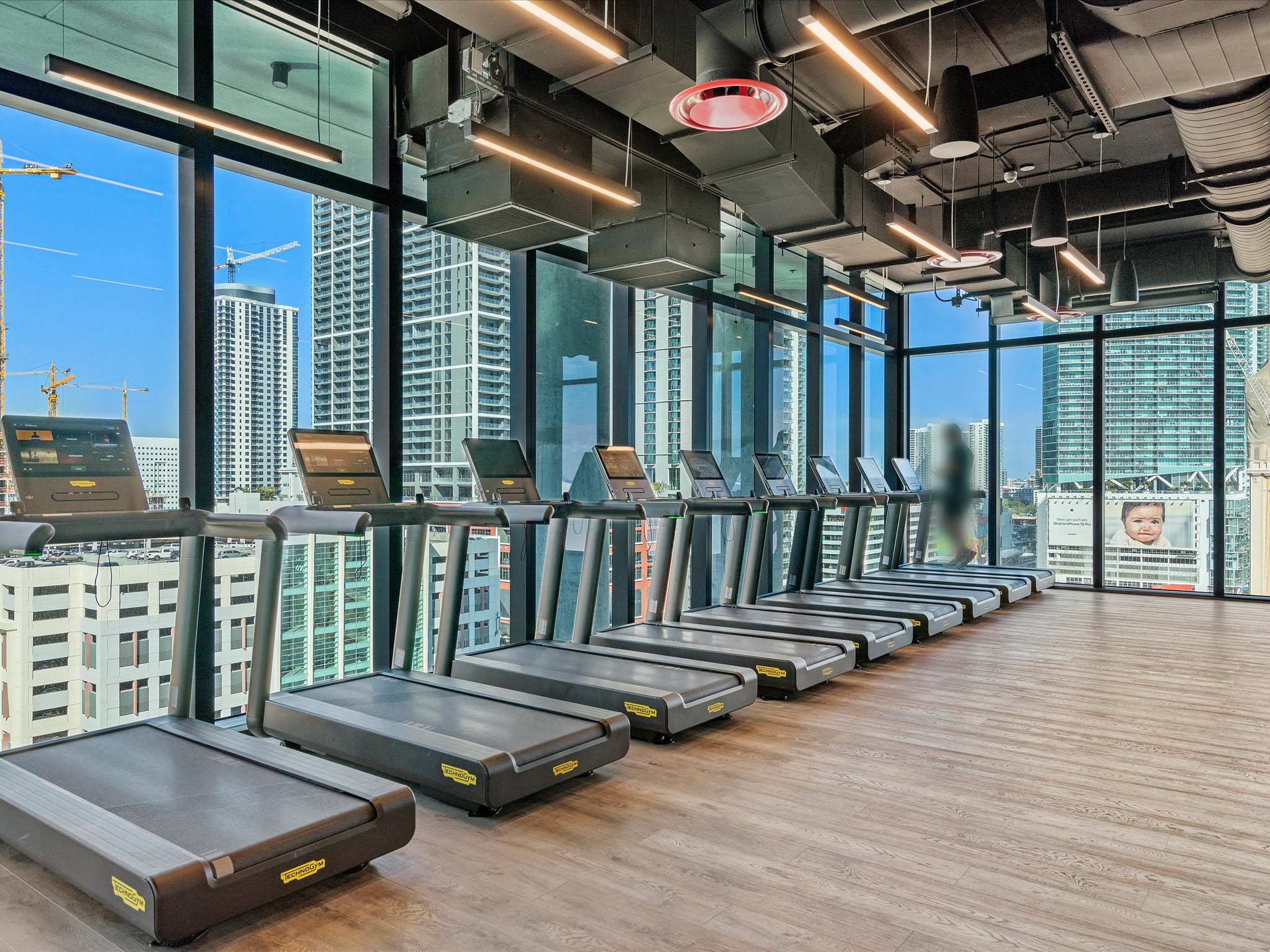 Modern gym area equipped with a row of treadmills facing large windows that provide panoramic city views. The space features an industrial ceiling design.