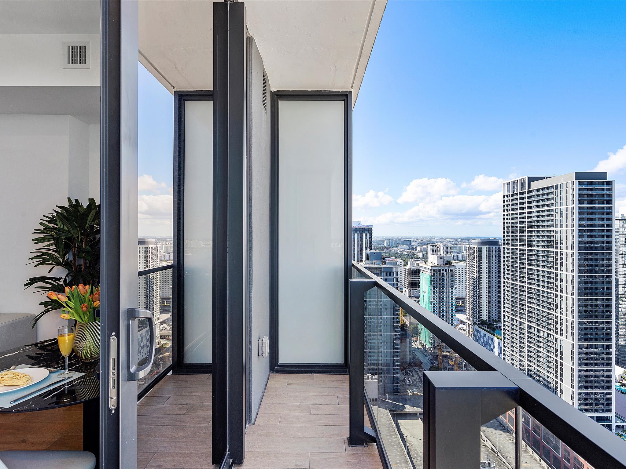 Private balcony offering expansive city views with numerous high-rise buildings under a clear blue sky. The balcony features a glass railing and a small outdoor dining setup with plants.