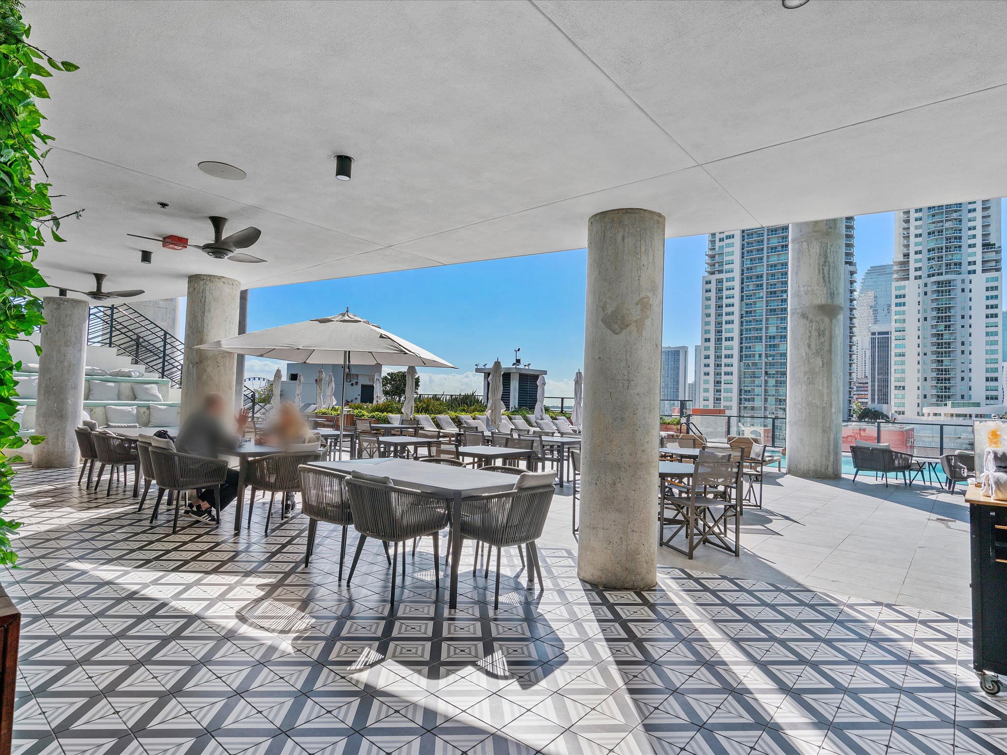 Covered outdoor patio dining area with multiple tables and modern chairs, featuring a distinctive patterned tile floor and large umbrellas for shade. The space offers views of surrounding city buildings.