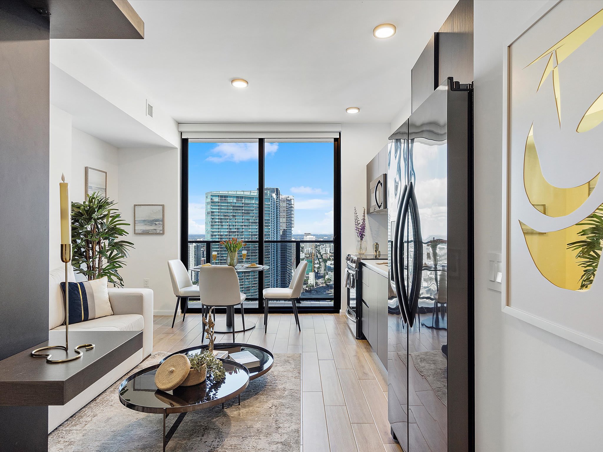 Open-concept living area showcasing a modern kitchen with dark cabinetry and stainless steel appliances, a small dining table, and a cozy living space with a sofa. Large windows provide a bright city and bay view.
