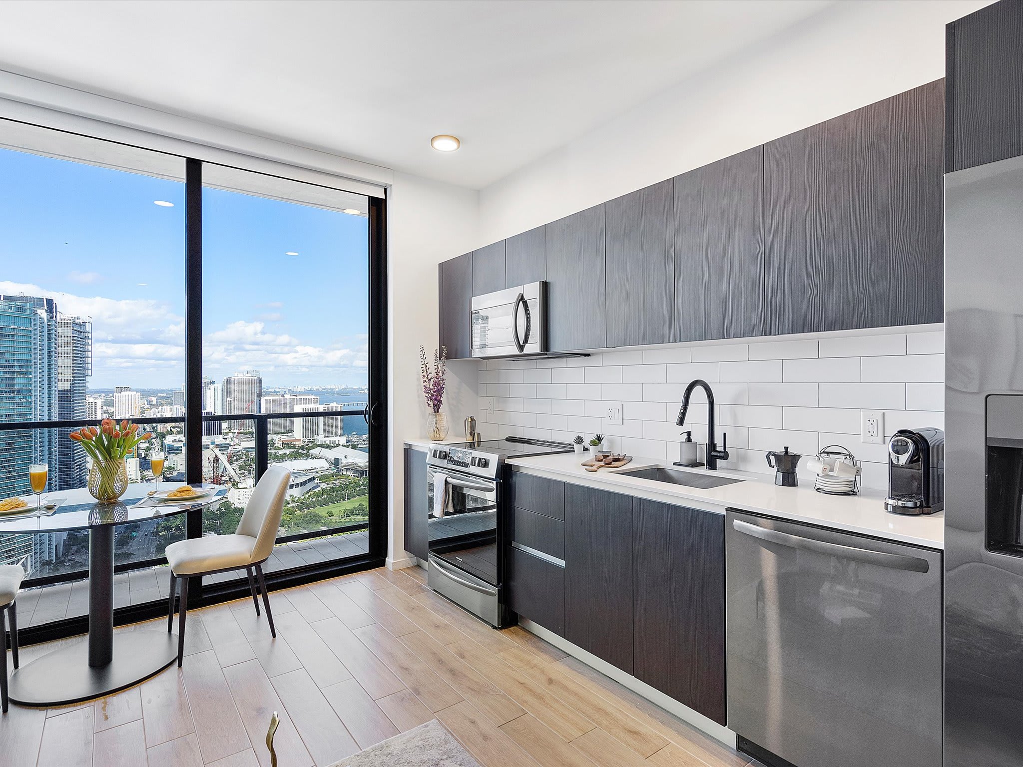 Modern kitchen featuring dark flat-panel cabinetry, white subway tile backsplash, stainless steel appliances including a stove, microwave, and dishwasher, and a black faucet over a sink. A small dining table with a city view is adjacent.