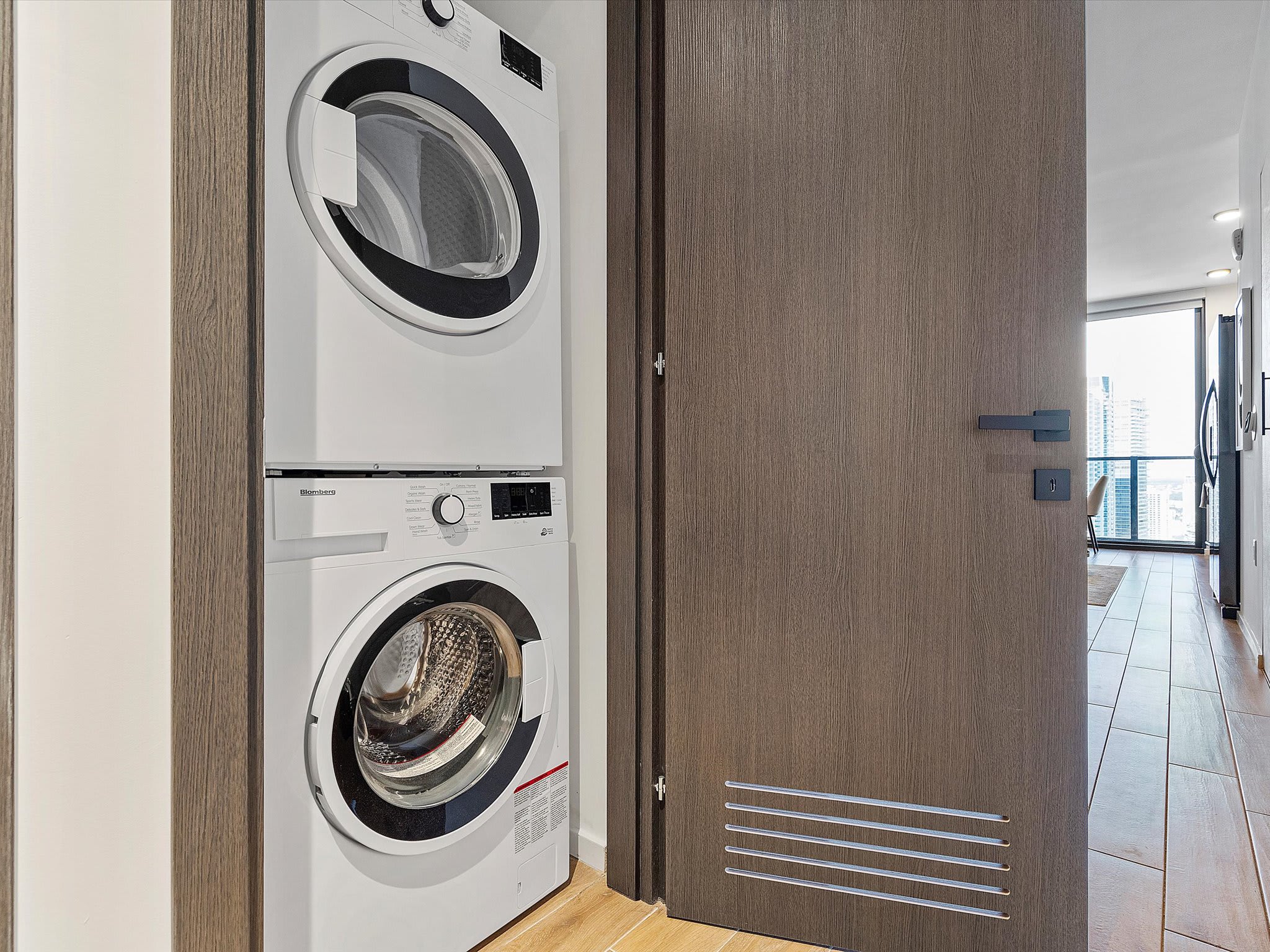 Stacked white washer and dryer units are neatly integrated into a utility nook with light wood flooring, adjacent to a dark wood door.