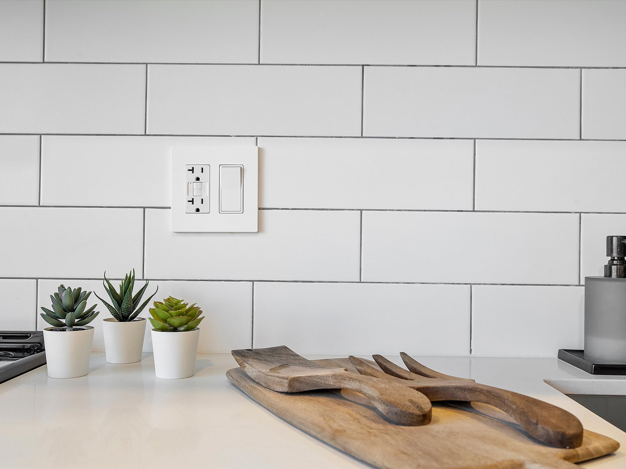 Close-up of a modern kitchen featuring white subway tile backsplash, a white electrical outlet with light switches, and small potted succulent plants on a white countertop. Wooden serving utensils are also visible.