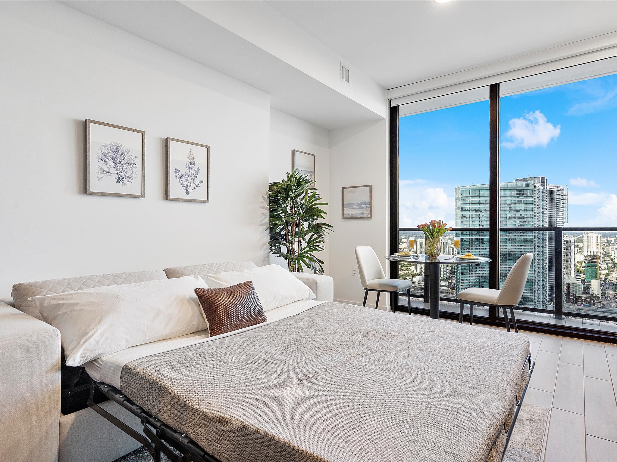 The studio's living area transformed into a sleeping space, with the light beige sofa converted into a pull-out bed. A small dining table is visible near the large windows, offering city views.