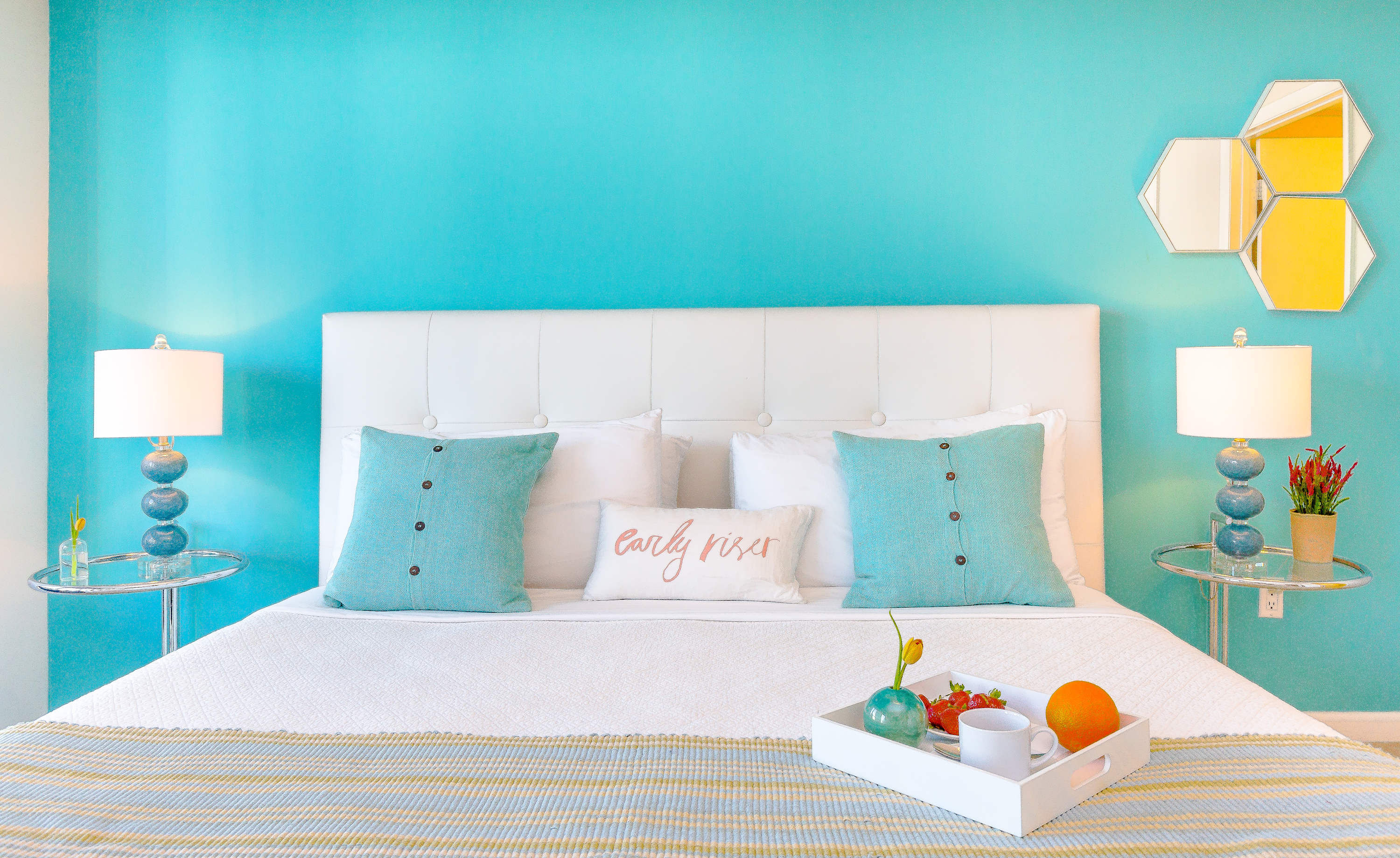 Bright bedroom with a white tufted headboard against a vibrant turquoise accent wall, featuring a king-sized bed with white bedding, teal pillows, and a breakfast tray.