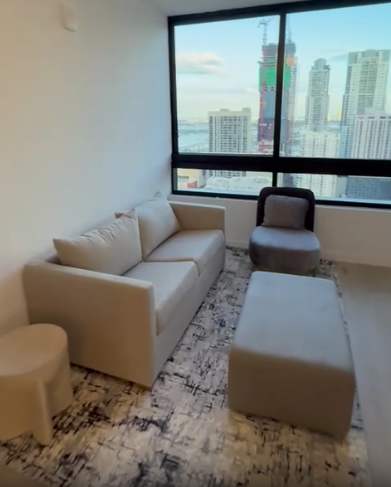 A modern living room featuring a light-colored sofa, a matching ottoman, and a single armchair, all arranged on a patterned rug. Large floor-to-ceiling windows offer panoramic city views of high-rise buildings and a body of water.
