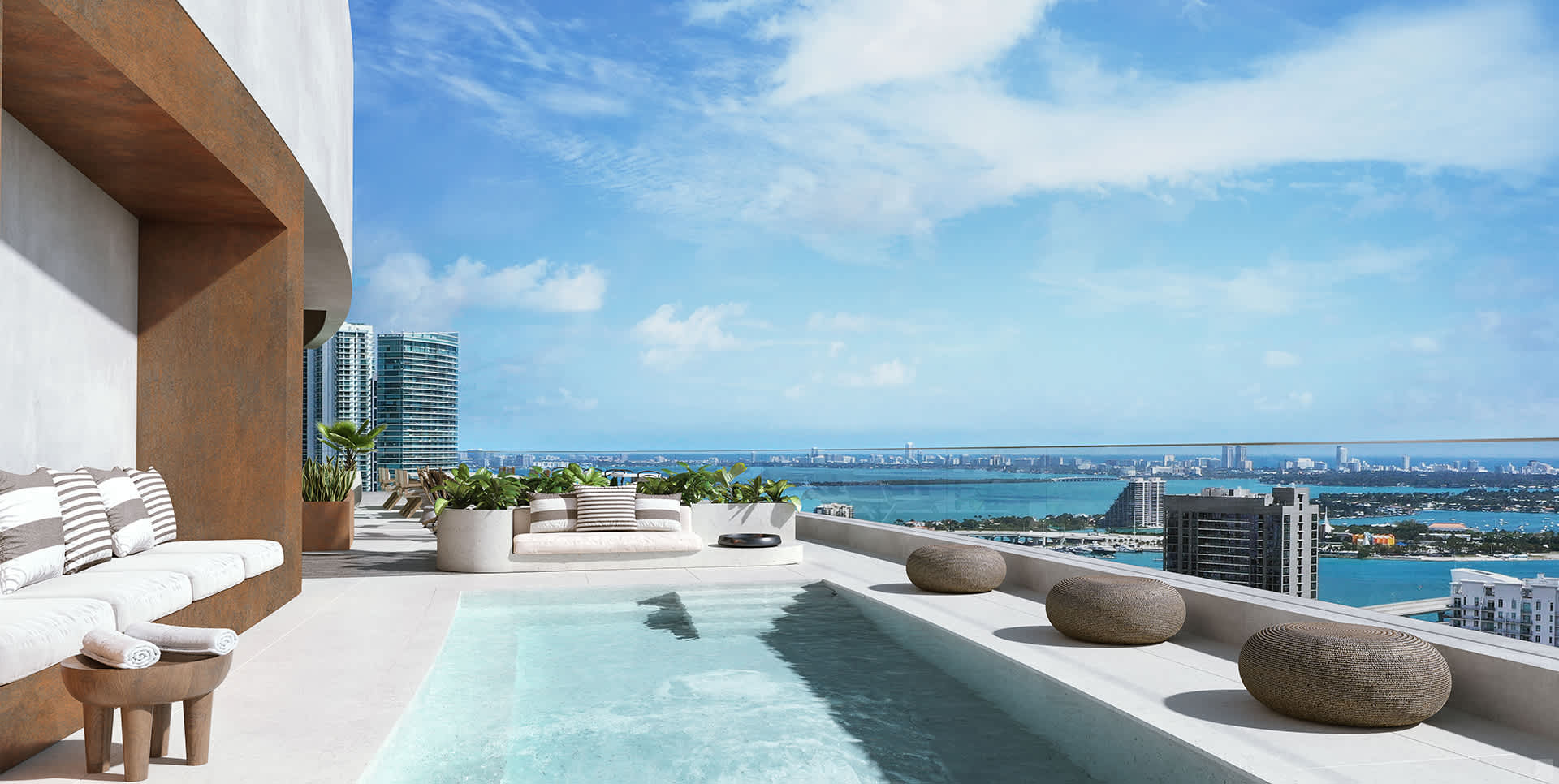 A luxurious rooftop terrace boasts a rectangular plunge pool, comfortable outdoor seating with striped cushions, and panoramic views of a city skyline and a vast bay under a clear blue sky.