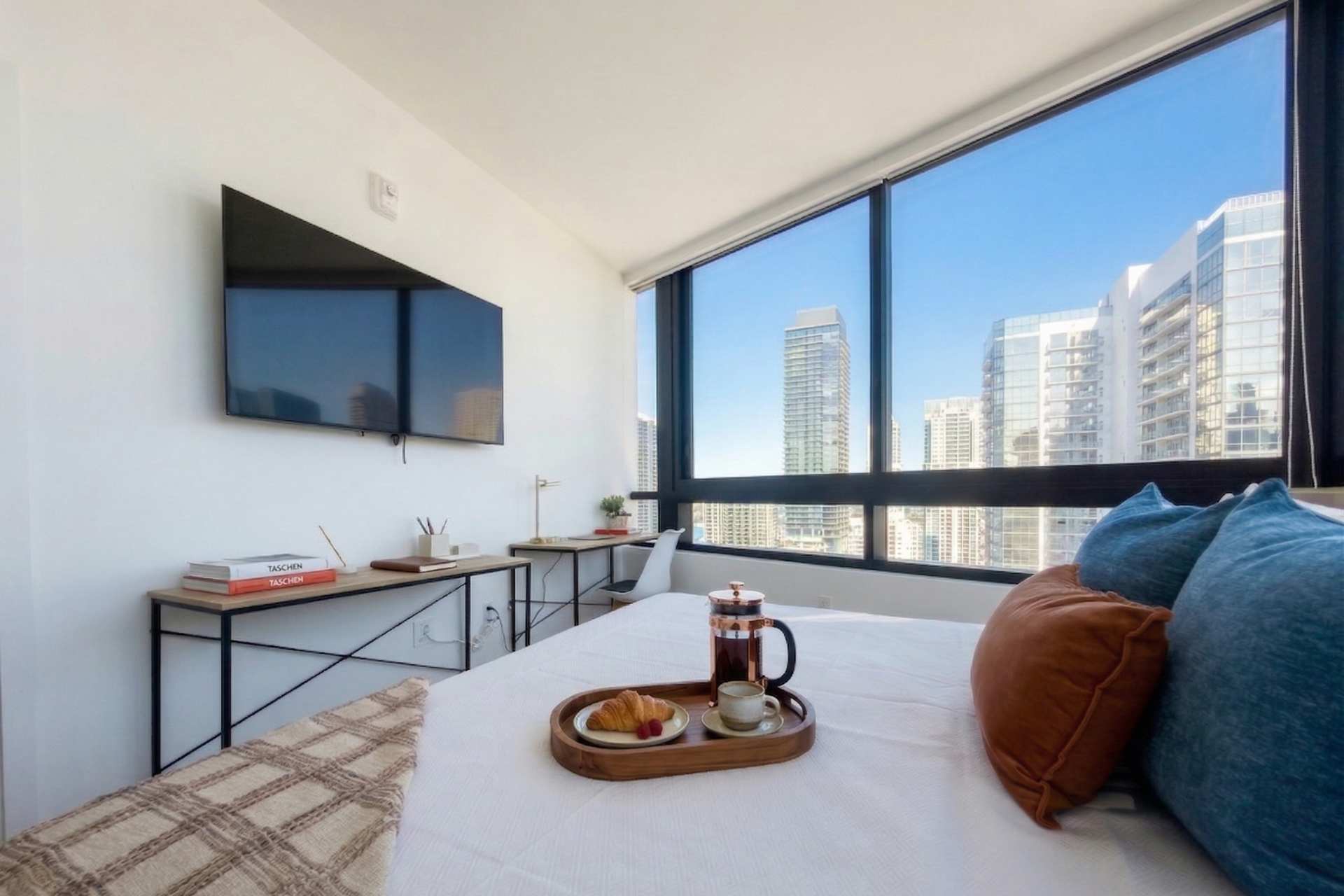 A modern bedroom with a bed featuring white linens and accent pillows, facing a wall-mounted TV. A minimalist desk and chair are placed by the large window, offering city views.