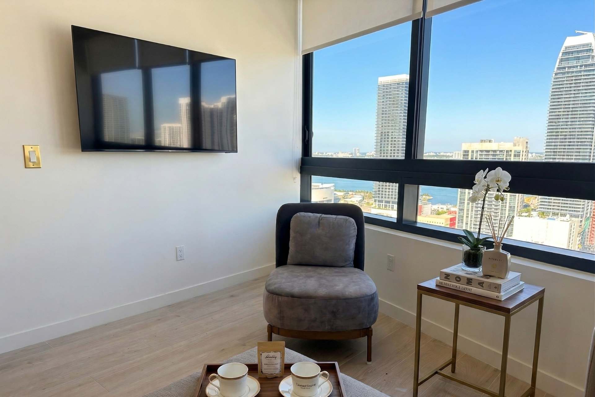 A comfortable grey accent chair sits by a large window, offering a partial city and water view. A small side table with decor and a wall-mounted TV complete the cozy corner.