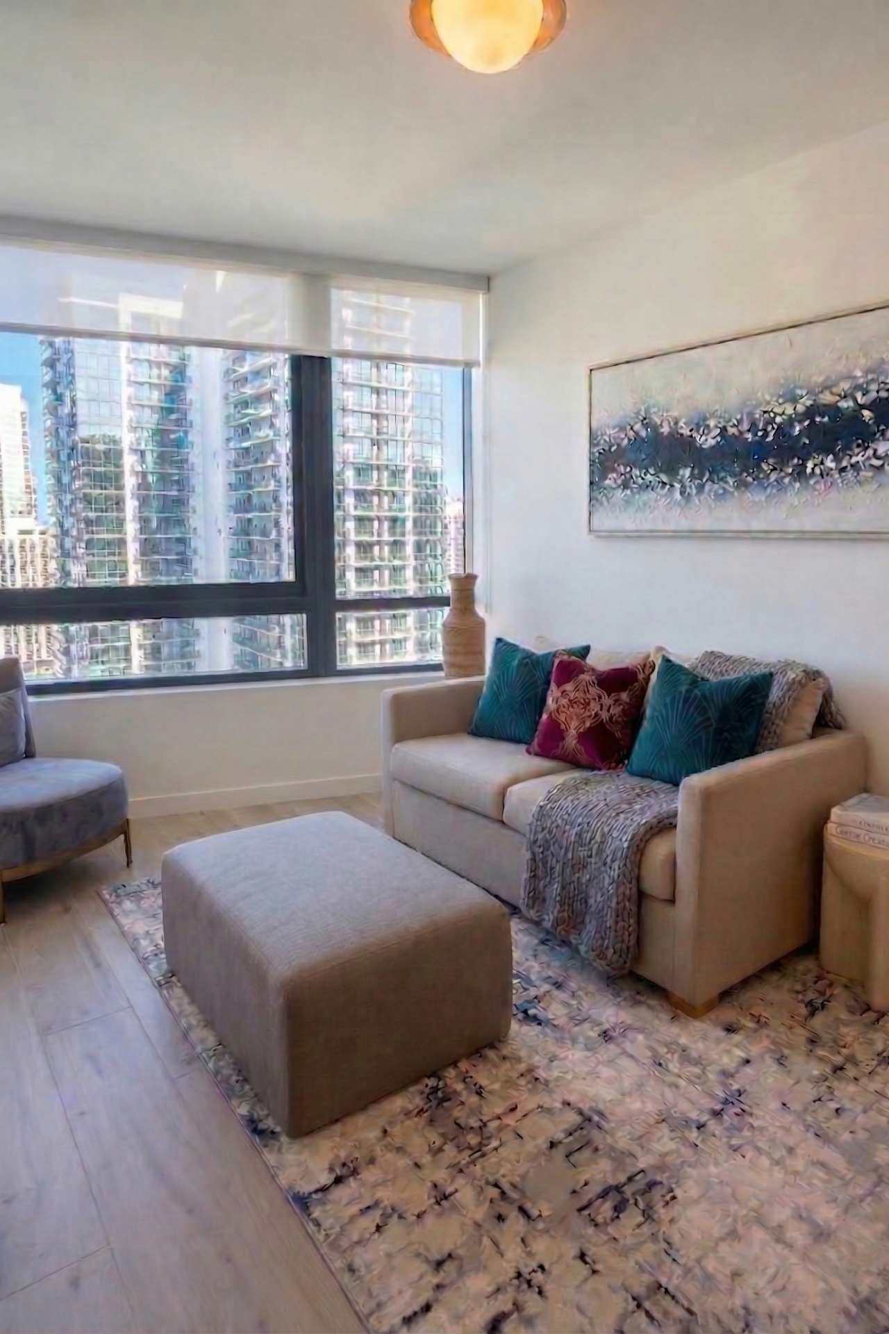 A cozy living room with a light-colored sofa adorned with colorful throw pillows, an ottoman, and a grey accent chair. Large windows offer city views, and an abstract painting hangs above the sofa.