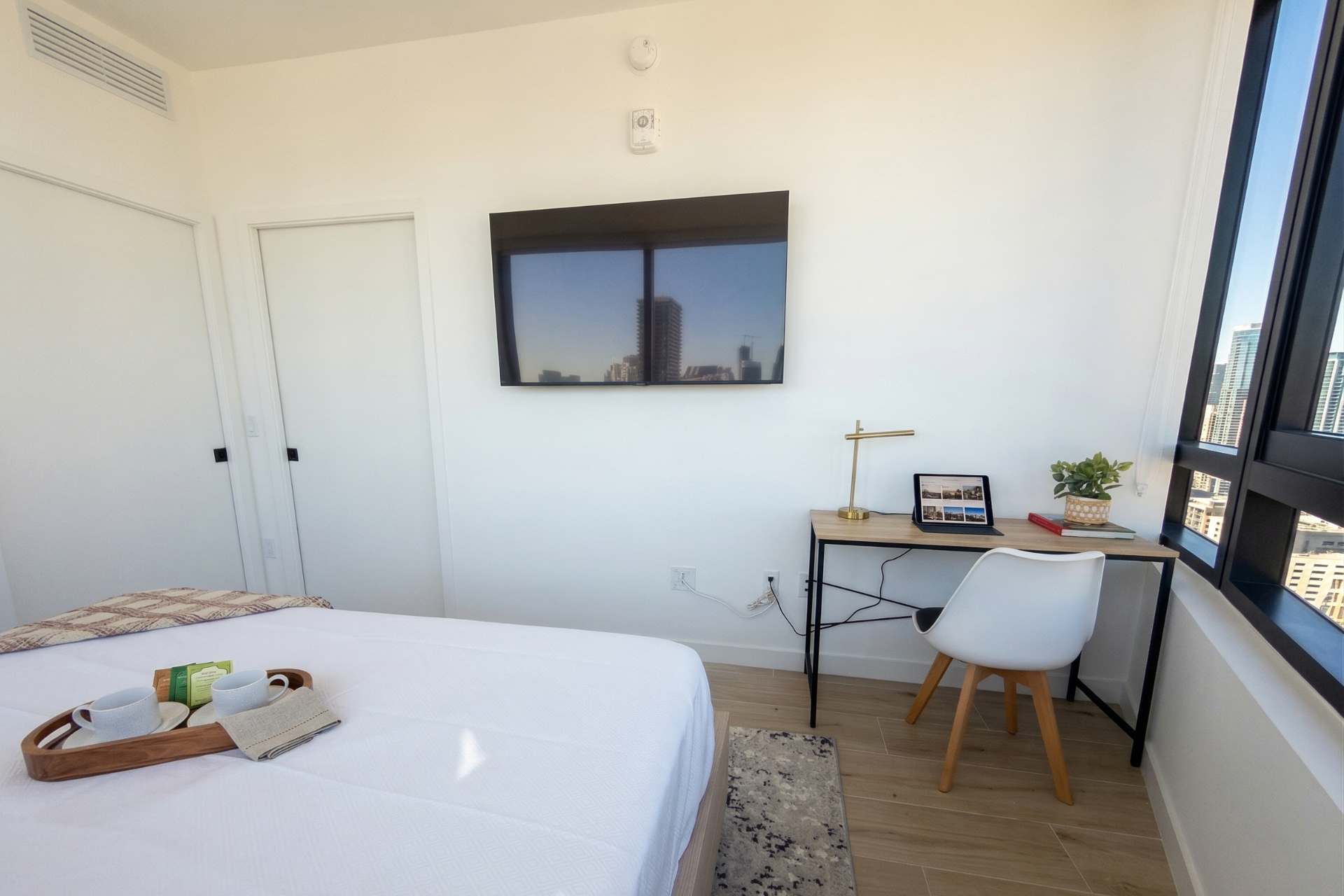 A bright bedroom with a bed, a wall-mounted TV, and a functional desk setup by the window. The room features light wood flooring and white walls, with city views outside.
