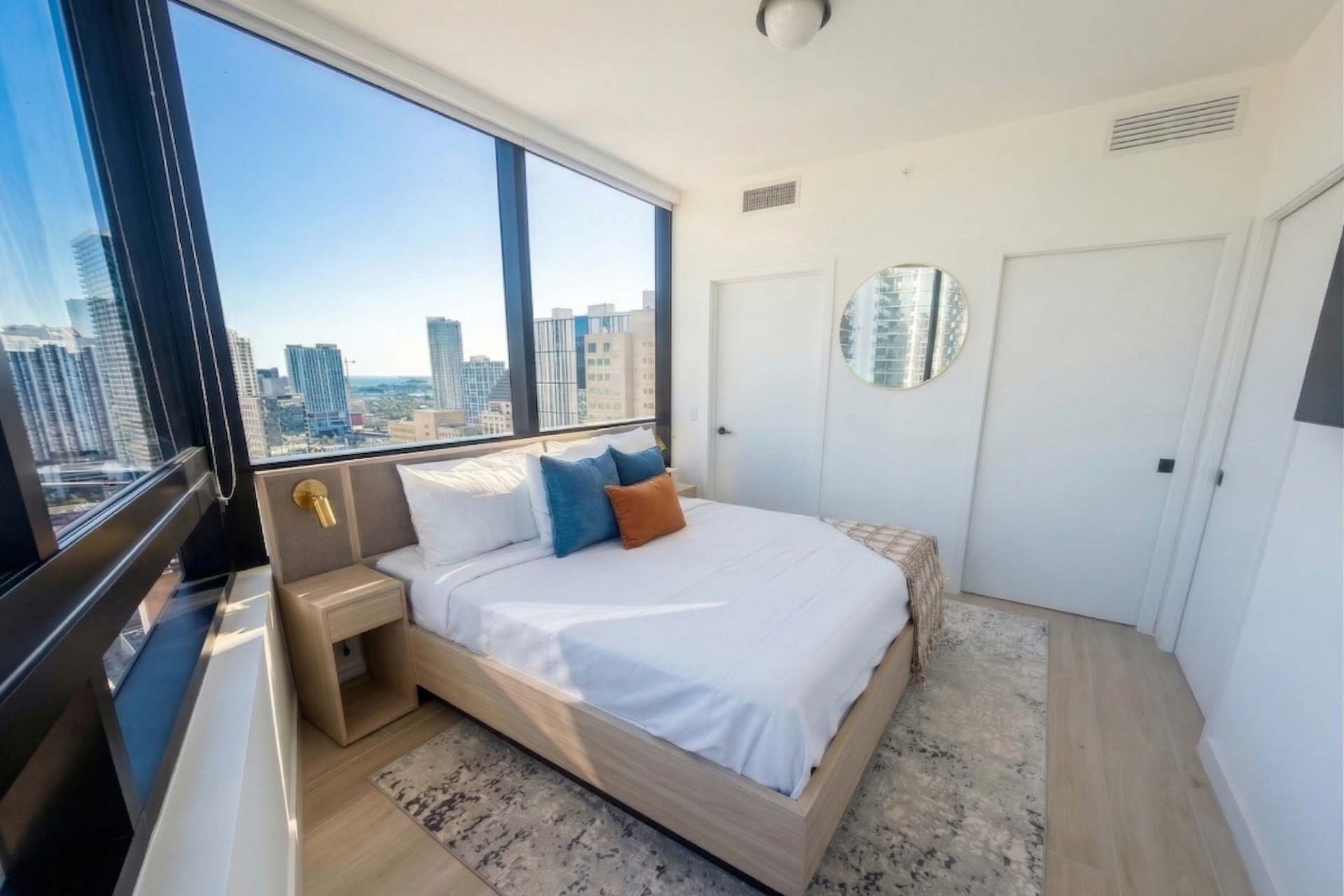 A bright bedroom featuring a light wood bed with white linens and colorful pillows, positioned against a wall with a round mirror. Large windows offer panoramic city views.